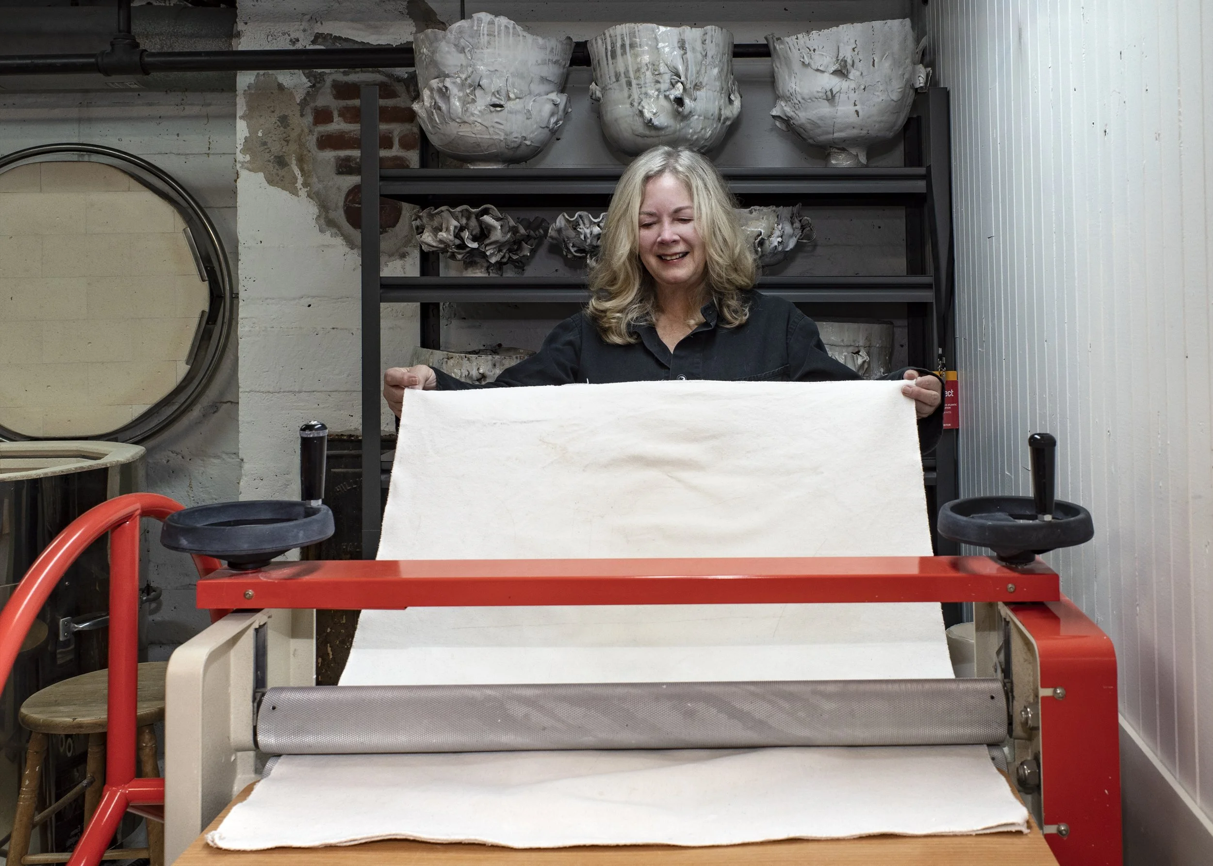 Woman operating a printing or textile machine in a workshop with shelves of large ceramic or plaster molds in the background.