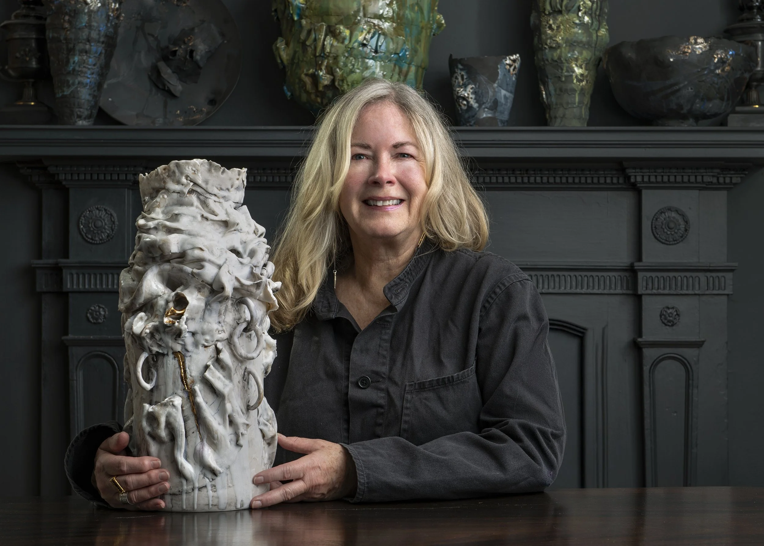 Heather is sitting at a dark wooden table holding a large, abstract, textured sculpture in a room with dark walls and a decorative dark fireplace in the background.