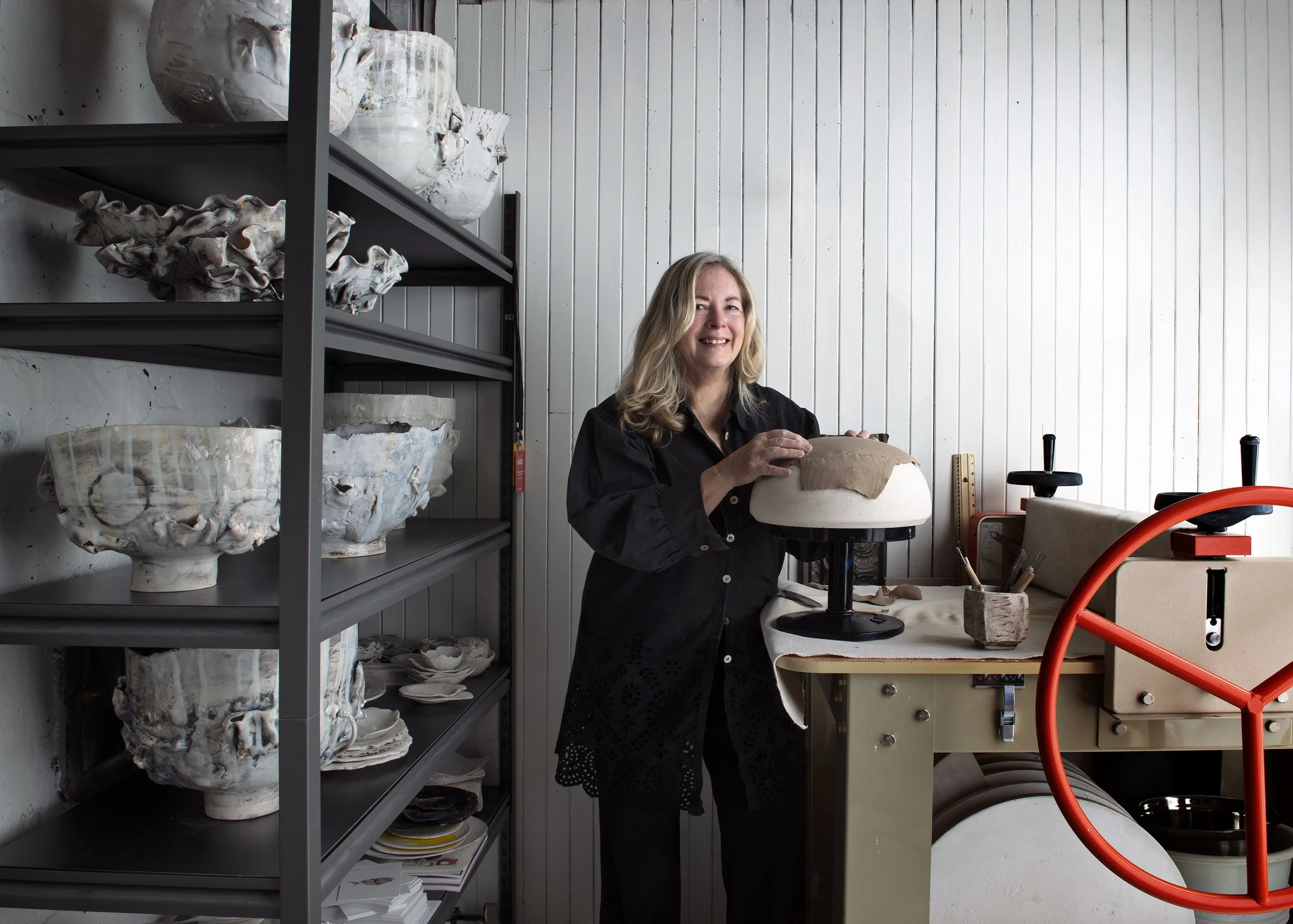 Heather is working on a pottery form in her studio, with shelves of ceramic pieces and white beadboard-panelled walls.