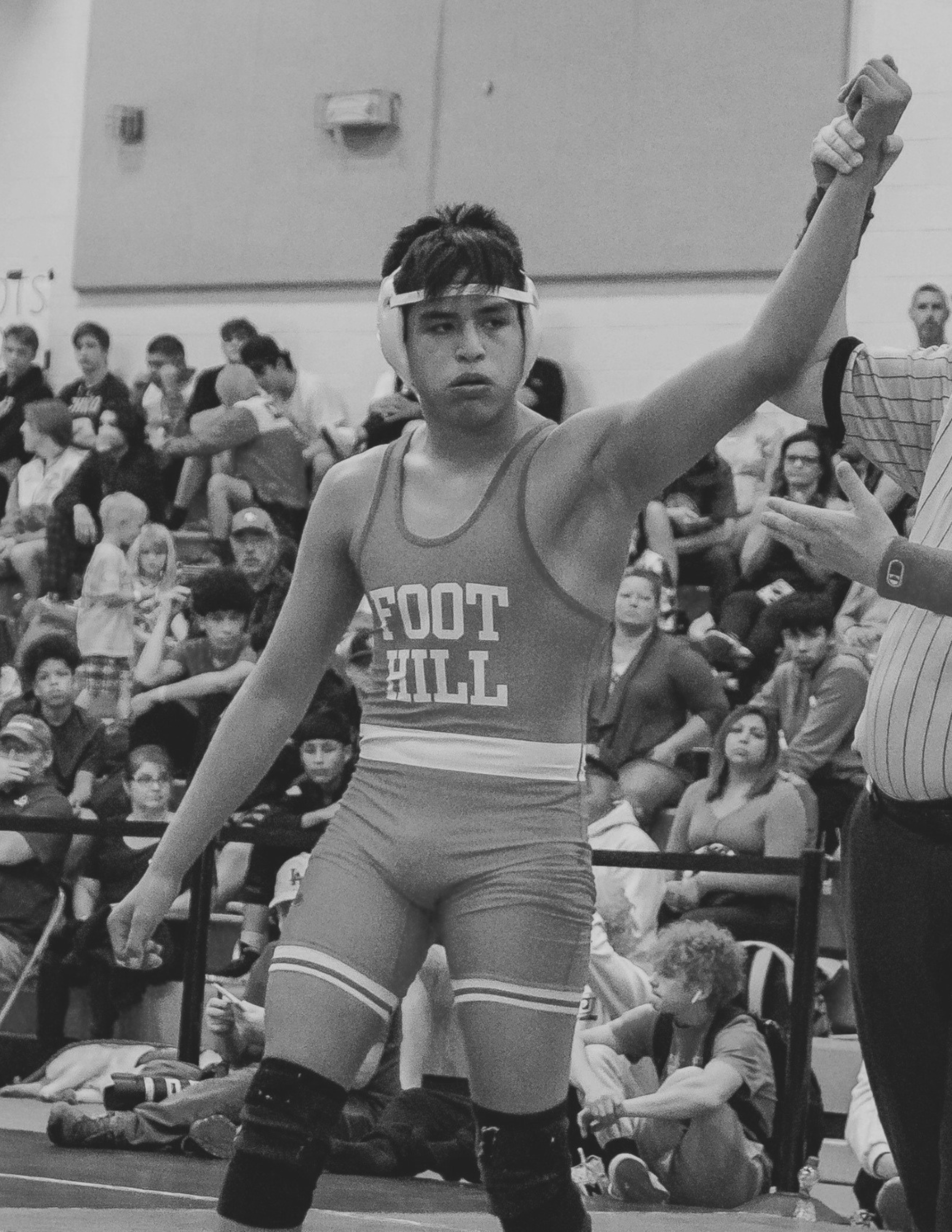 A young Shasta County, male wrestler in singlet with arm raised in victory in a gym, with an audience of children and adults sitting on the floor and standing in the background in Redding, CA