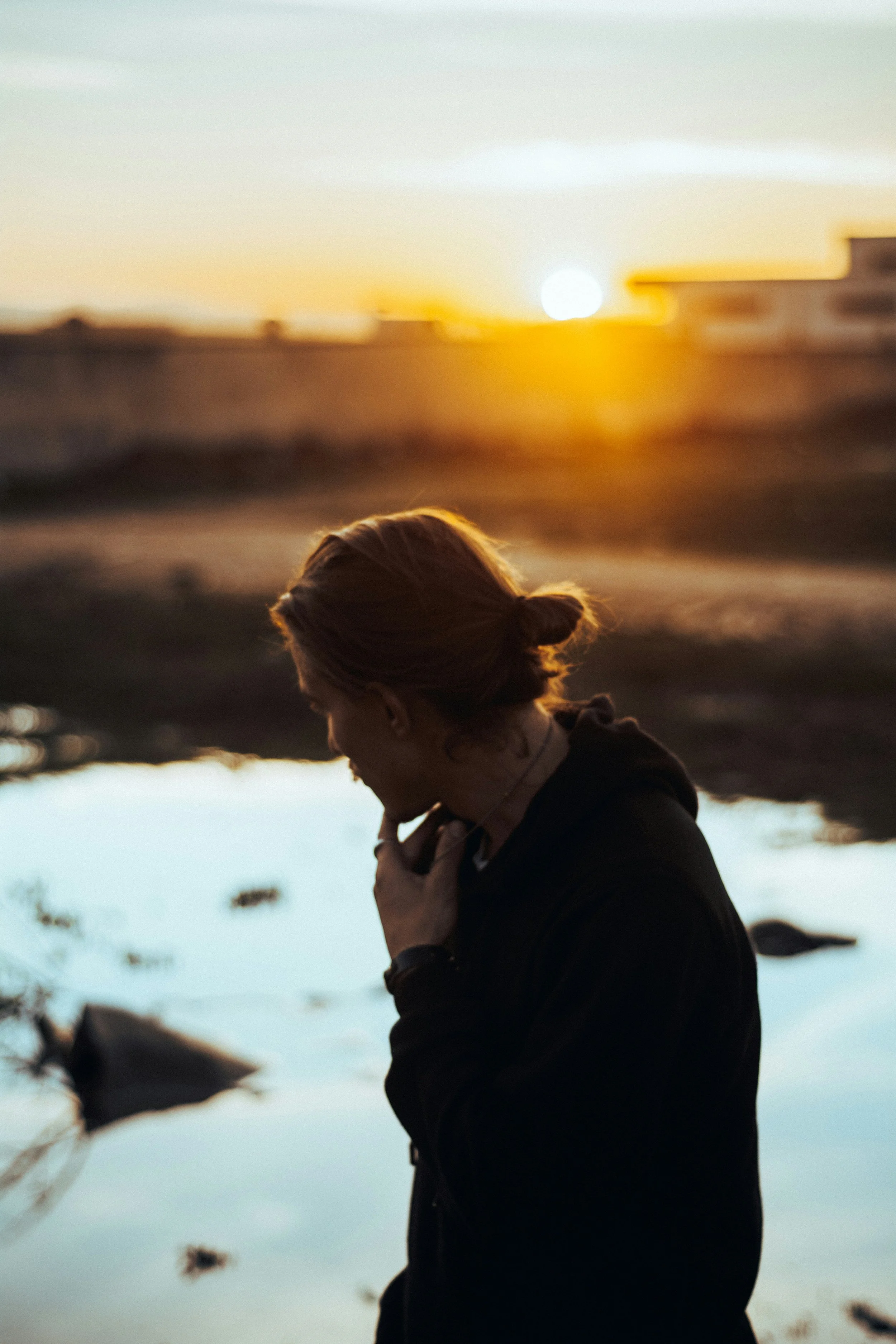 A woman stands alone outdoors at sunset, head bowed in quiet reflection, warm golden light behind her.