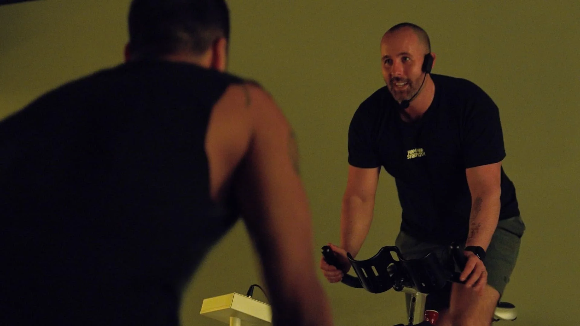 A fitness instructor giving a workout class to an individual on stationary bike, both are in a room with green walls.