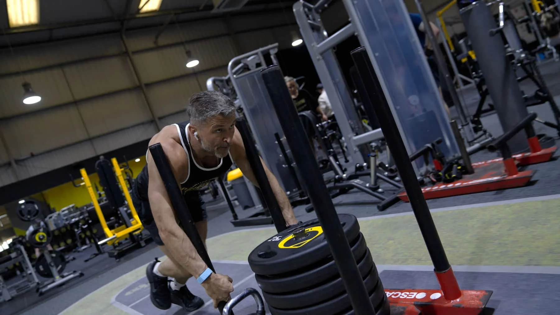 An older man with gray hair and a beard working out in a gym, pushing a sled loaded with weight plates. He is wearing a black vest, black shorts, black sneakers, and listening to wireless earbuds.
