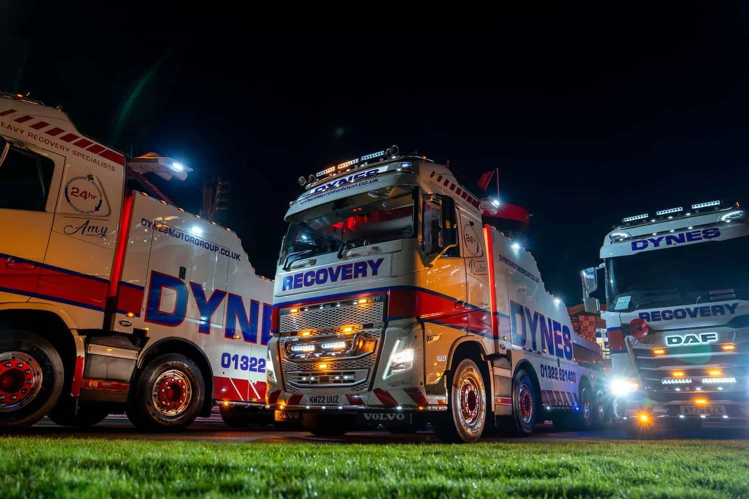 Dynes Motor Group recovery trucks lined up on the pit lane at Brands Hatch at night, with vehicle lights illuminated before the fireworks display