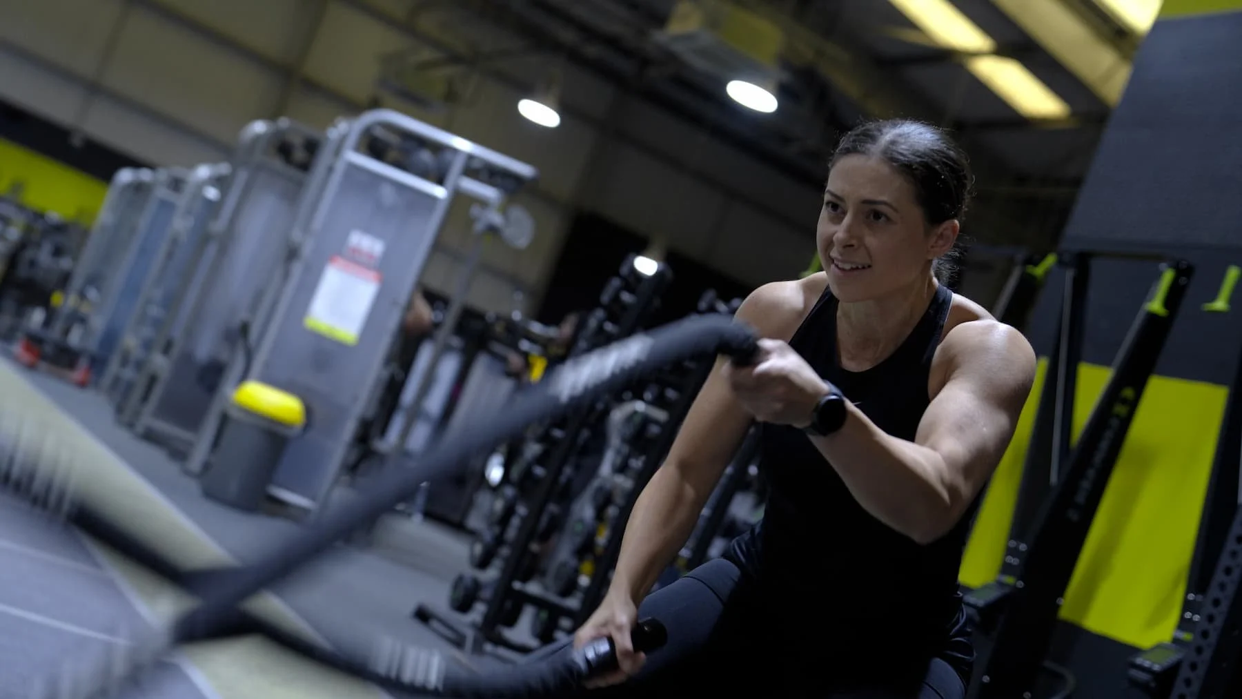 A woman working out with battle ropes at a gym, wearing a black sports tank top and black workout pants, with exercise equipment and yellow accents in the background.