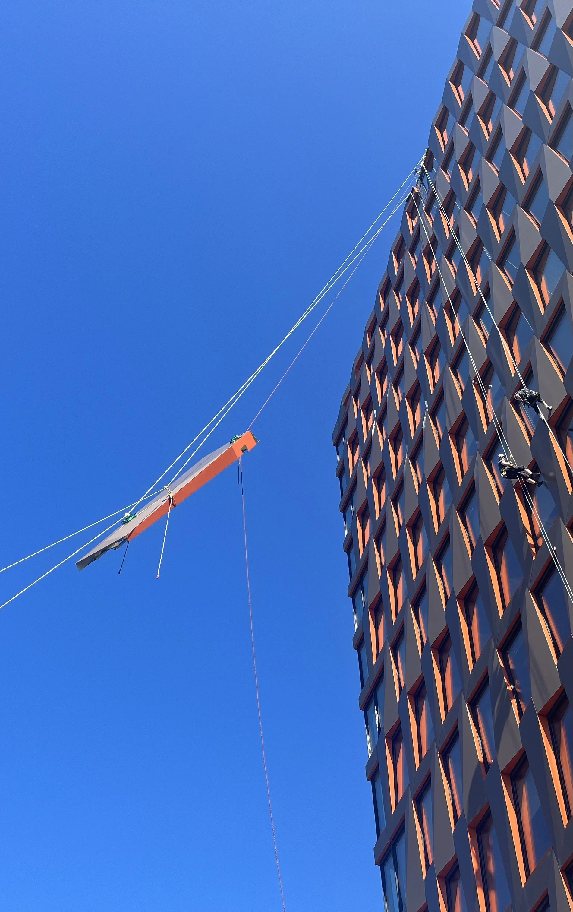 A facade element hanging in the air with rope access workers climbing on a building