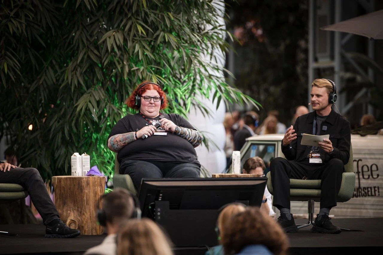 Two speakers, a woman with red hair and glasses, and a man with blond hair, sitting on chairs on a stage at a public event, wearing headphones. The woman is holding a microphone, and the man is holding papers. There are people in the audience, some wearing headphones, and a large leafy plant behind them.