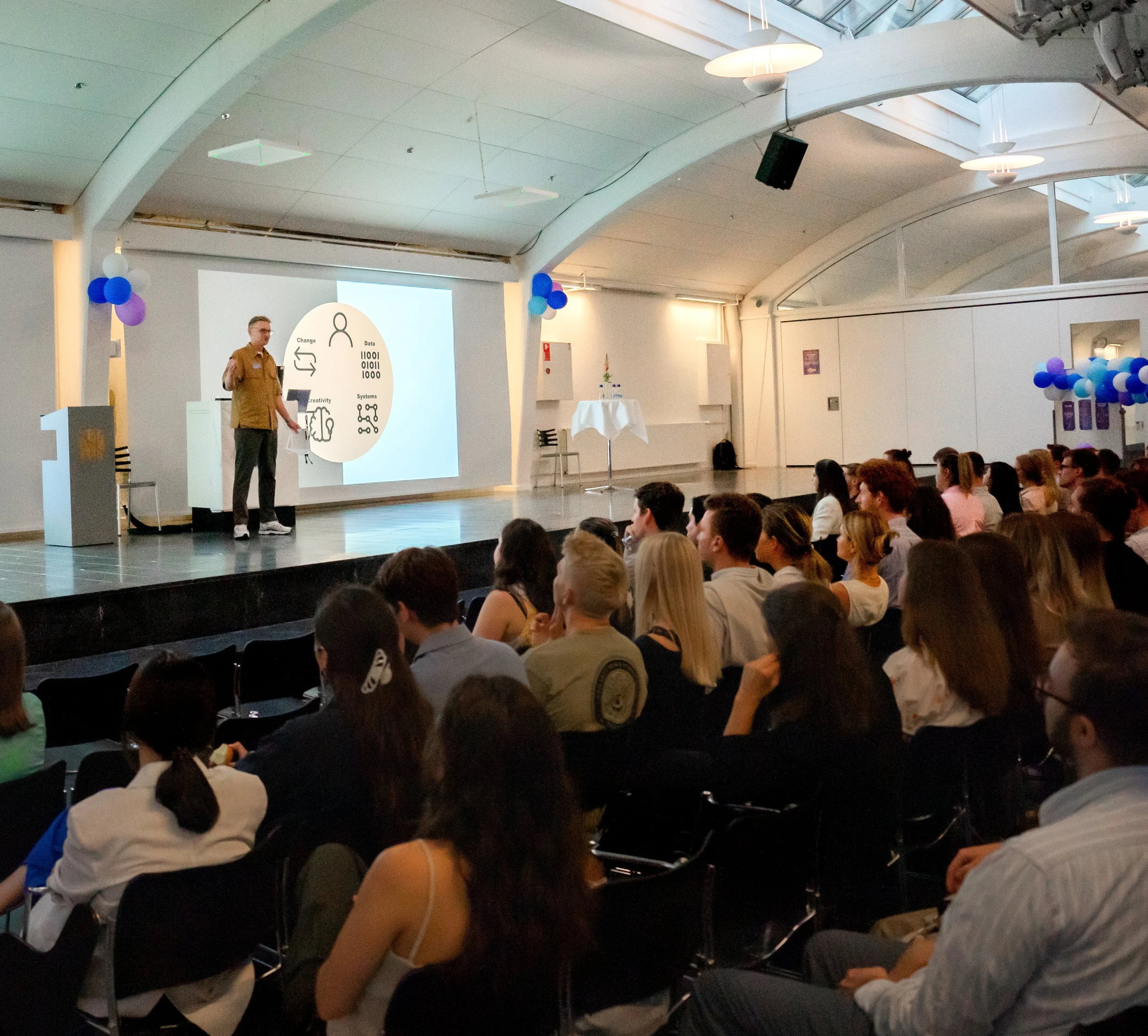 Speaker giving a presentation to an audience in a bright conference room with balloons.