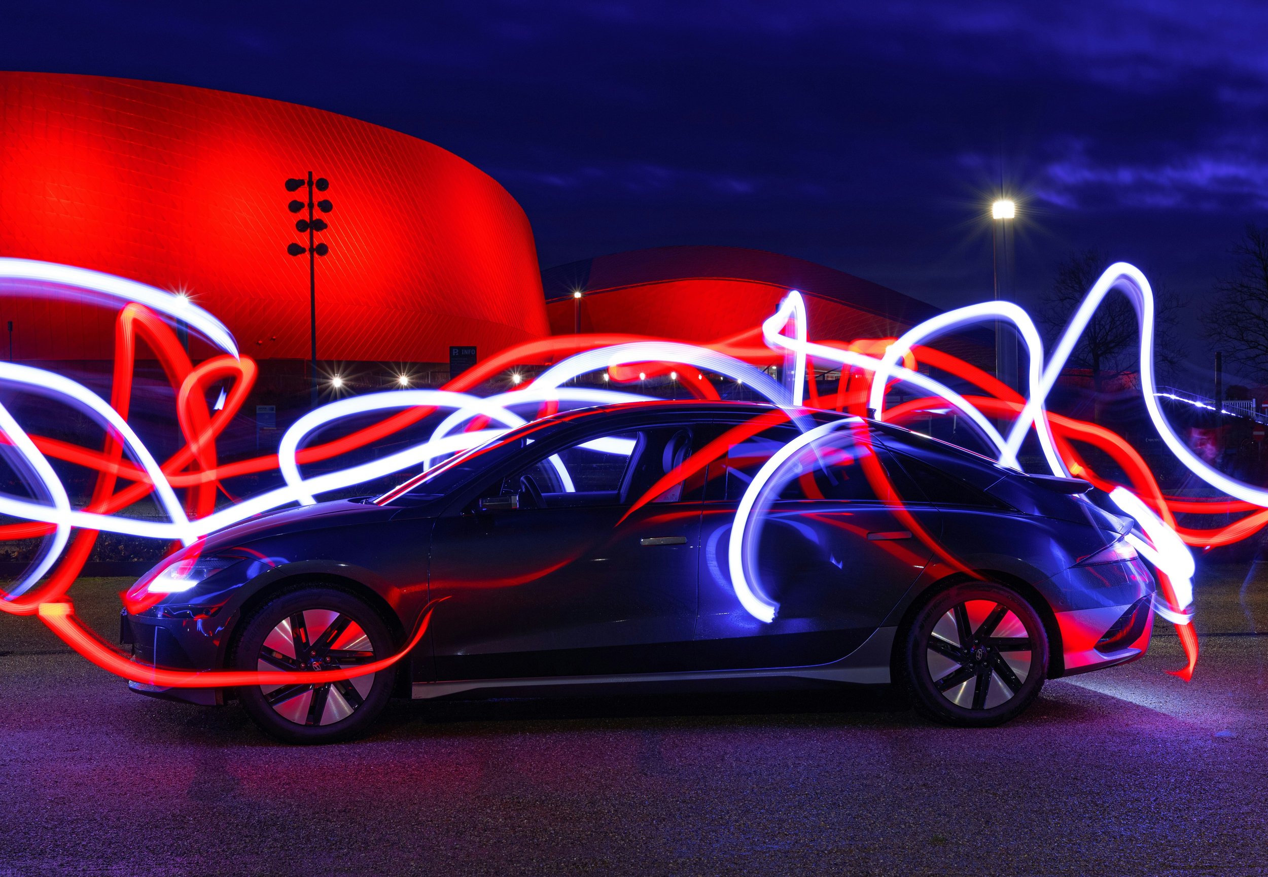Long exposure photo of a black car with red and white light streaks swirling around it, taken at night with a red building and streetlights in the background.