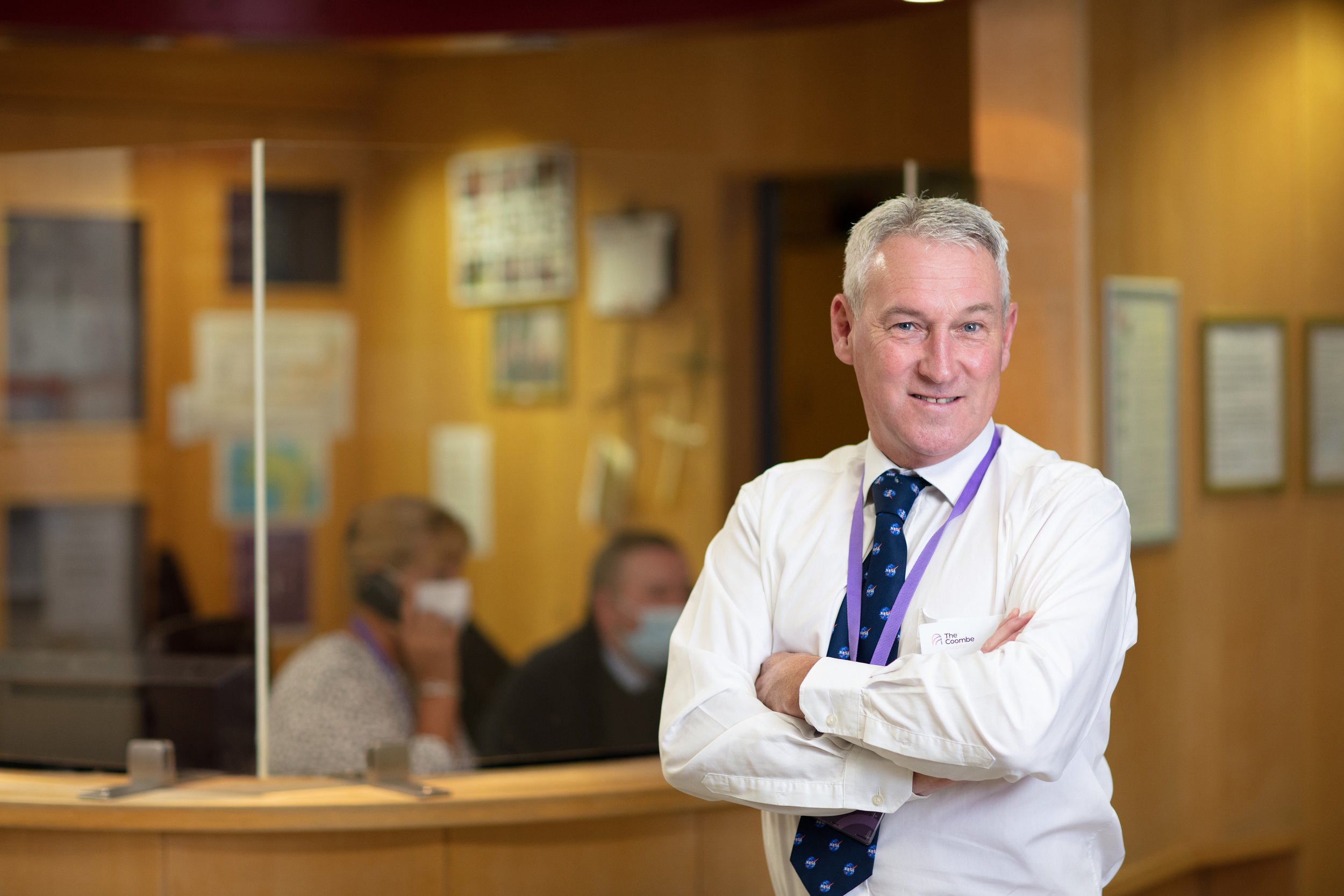 Man in business attire standing with arms crossed in a professional office setting, with people working behind a glass partition in the background.