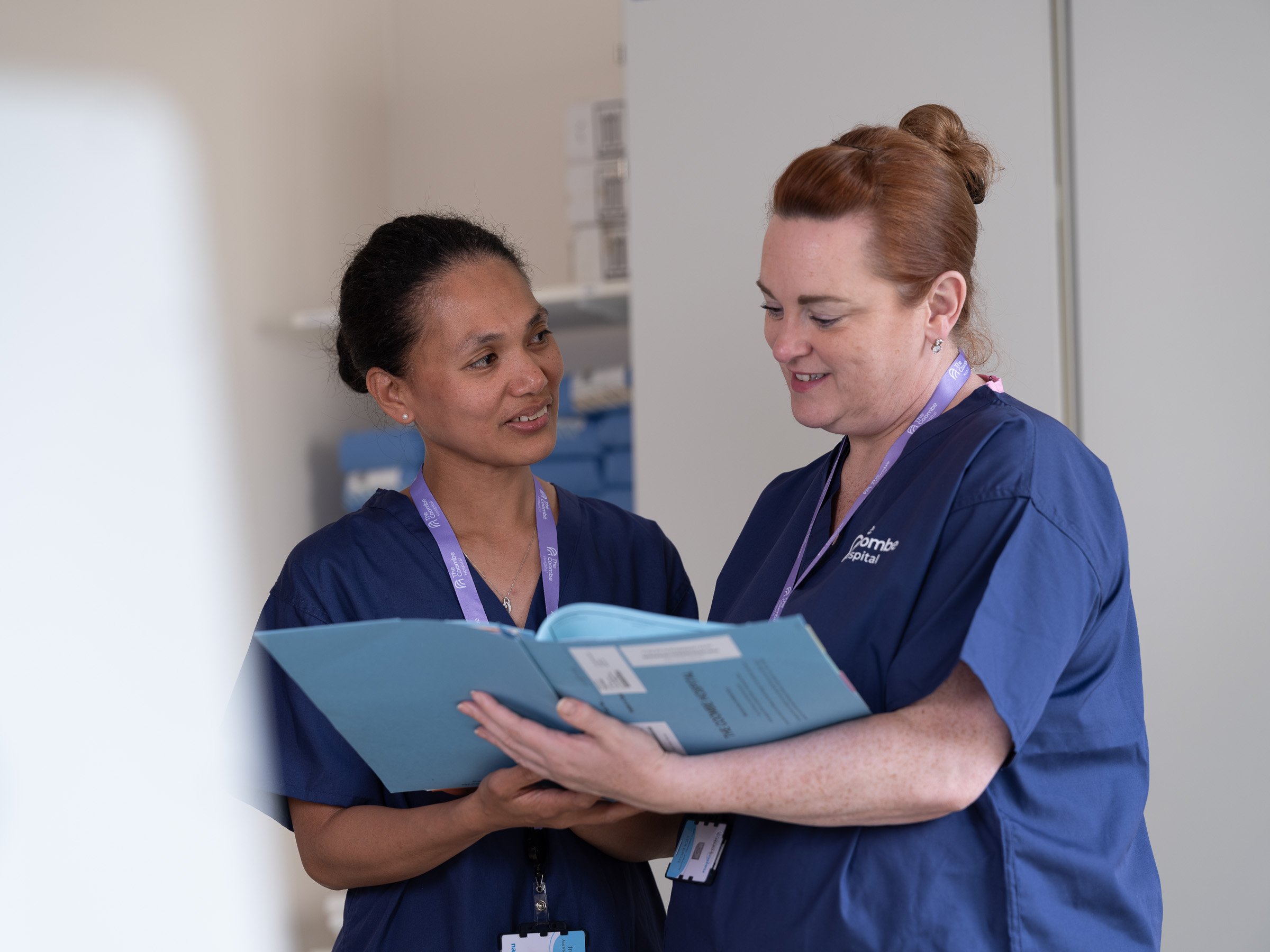 Two healthcare professionals in blue scrubs reviewing a file together in a clinical setting.