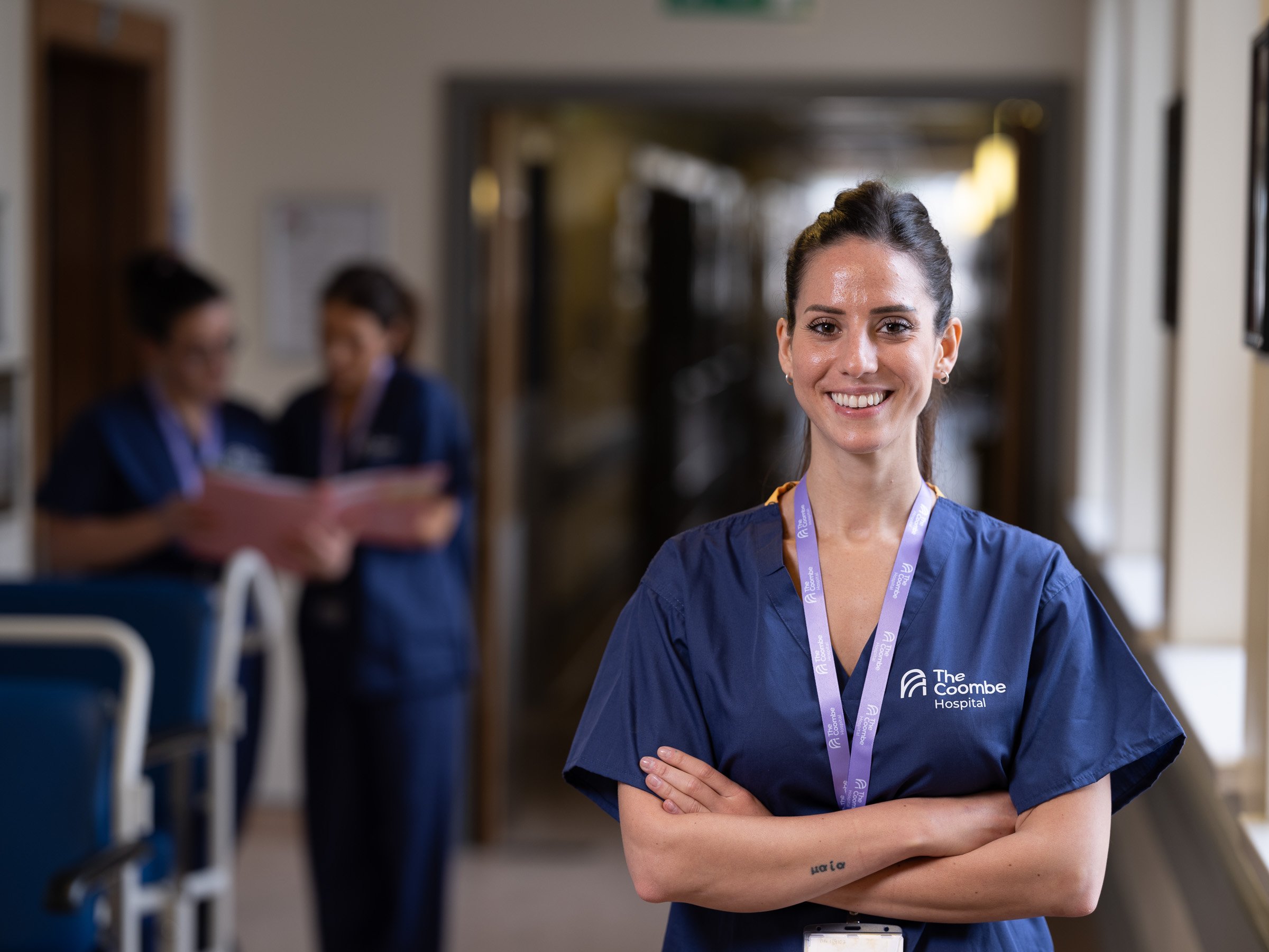A healthcare professional in blue scrubs with a lanyard, standing confidently in a hospital corridor with two colleagues in the background.