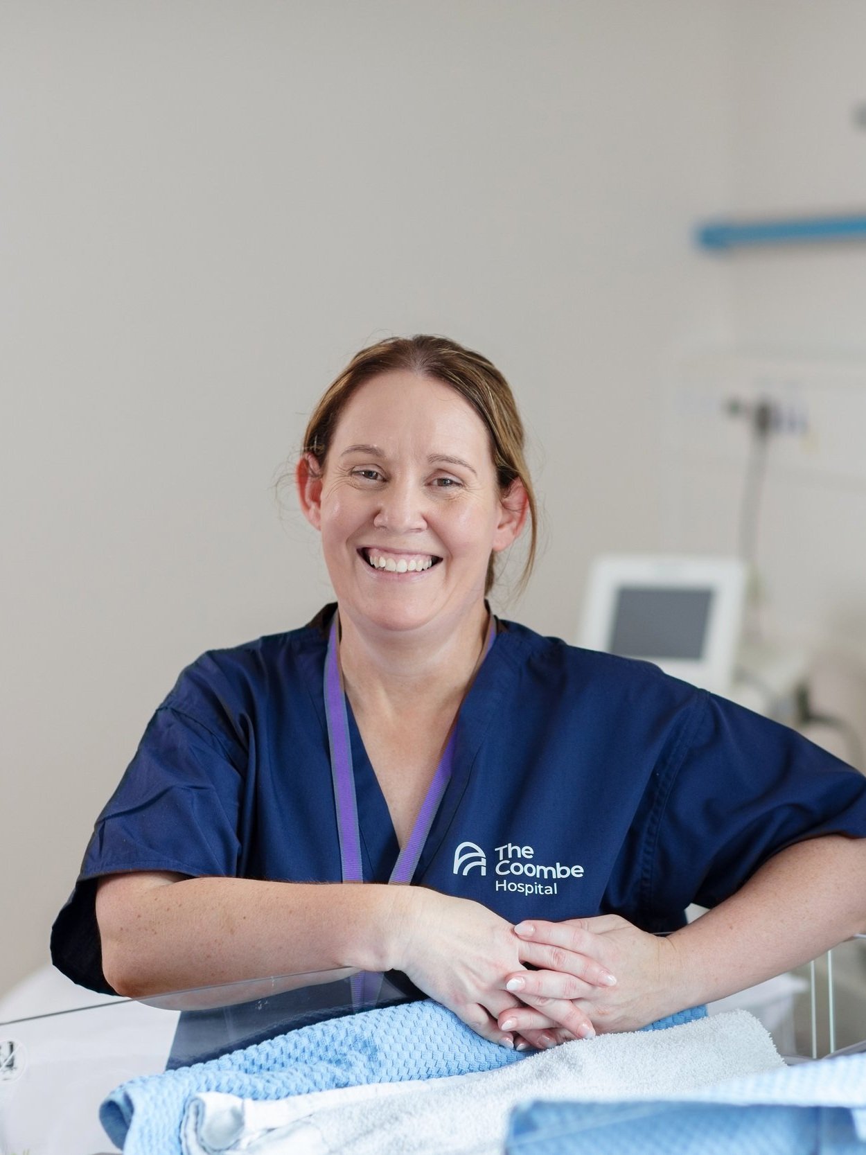 A smiling healthcare worker wearing a navy uniform with "The Coombe Hospital" logo, standing beside a blue and white blanket in a medical setting.