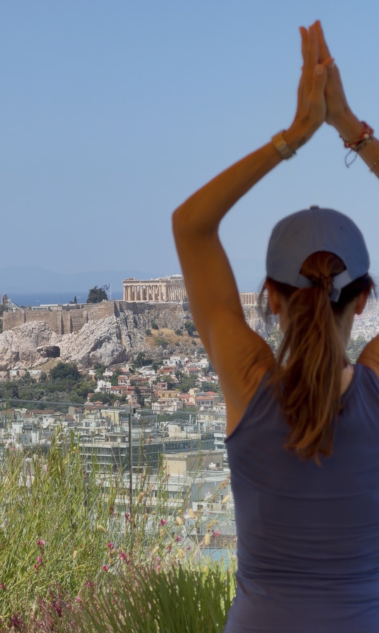 A person with tied-back hair wearing a gray cap stretches their arms upward in front of an ancient Greek temple on a hill, with a cityscape below under a clear blue sky.