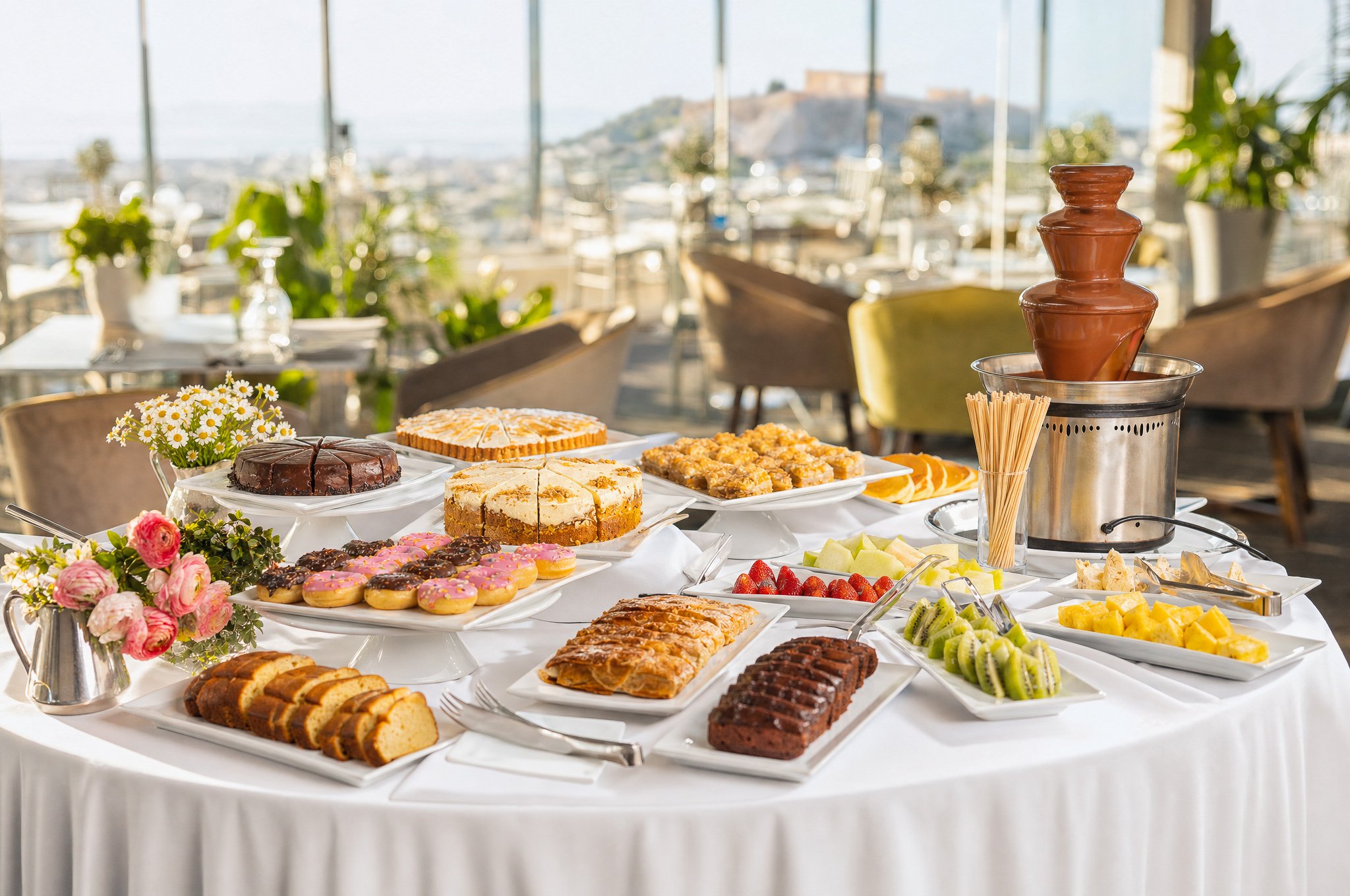 A table set with various desserts including cakes, donuts, and cookies, with flowers and a chocolate fountain in the background, in a bright room with large windows overlooking a cityscape.