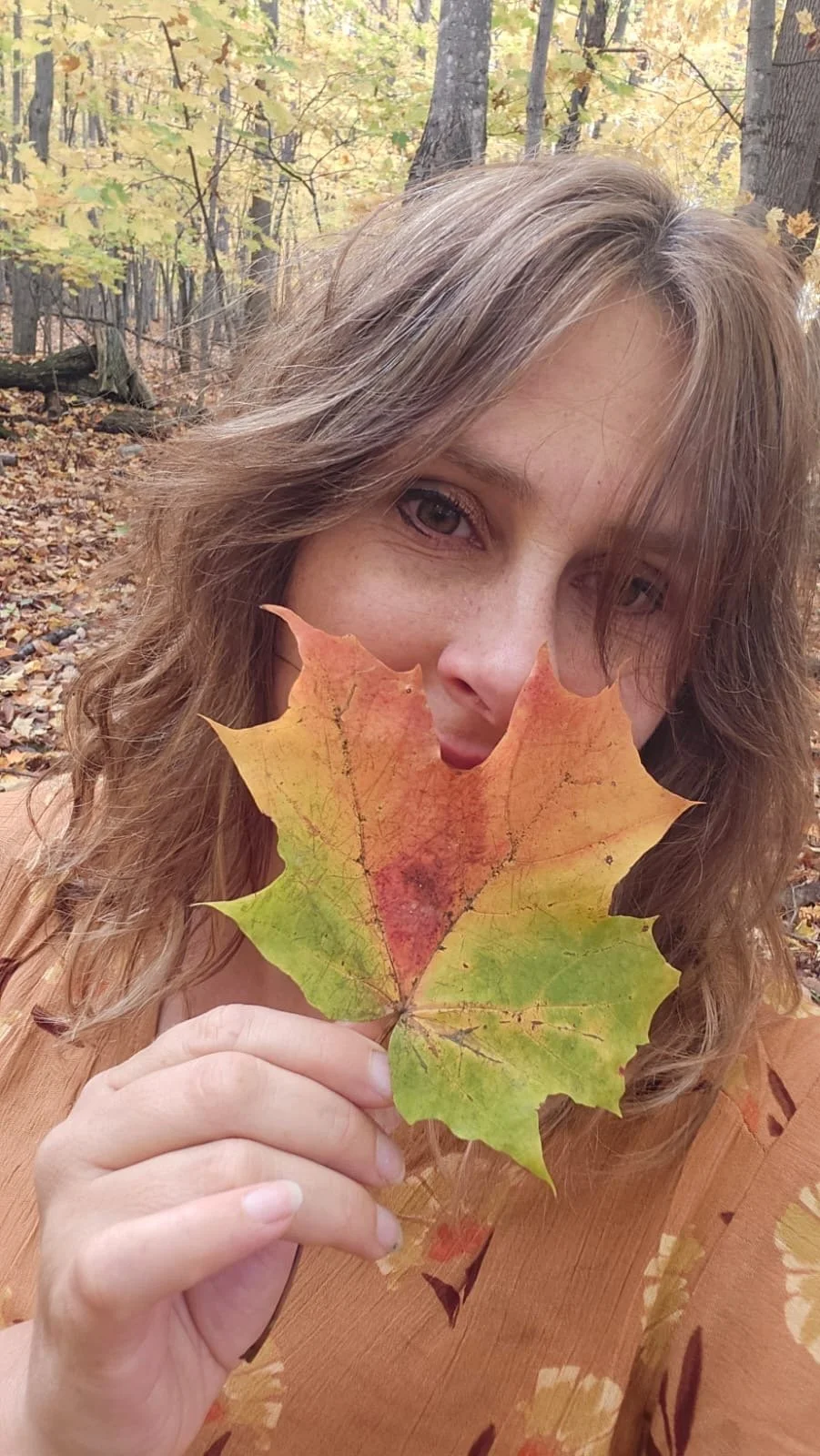A woman holding a multicolored autumn leaf in front of her face, partially covering her mouth, with a forest background of yellow and green leaves.