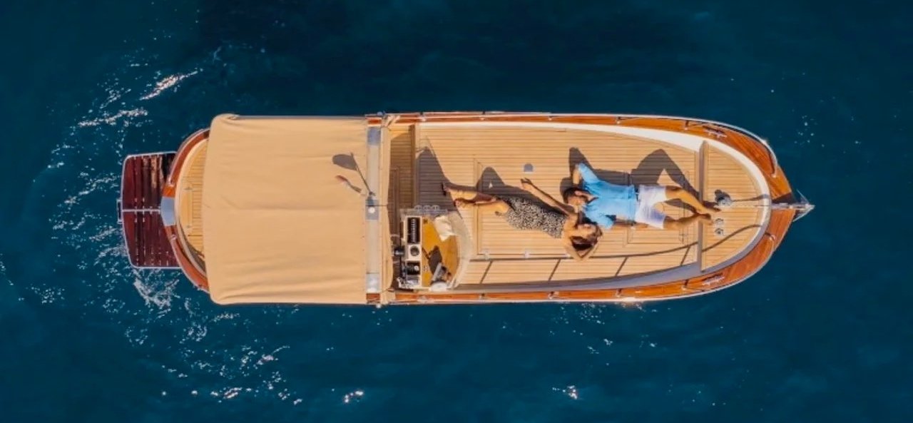 A boat with two people lounging on the deck, floating on blue water, seen from above.