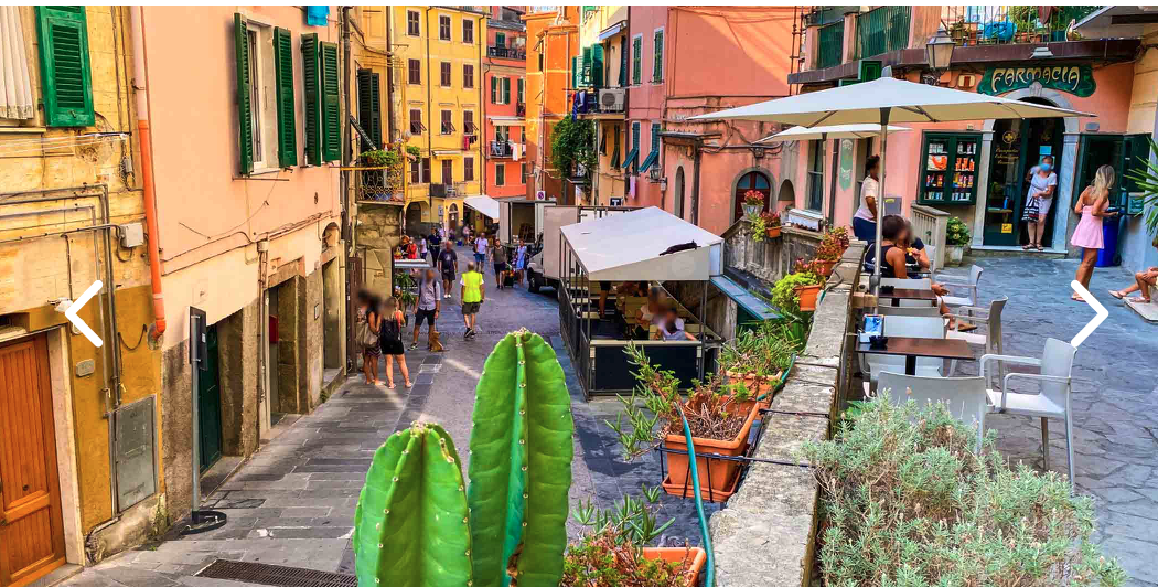 Vernazza with colorful buildings line a narrow street with outdoor cafes, potted plants, and people walking and sitting outside in a lively European town.