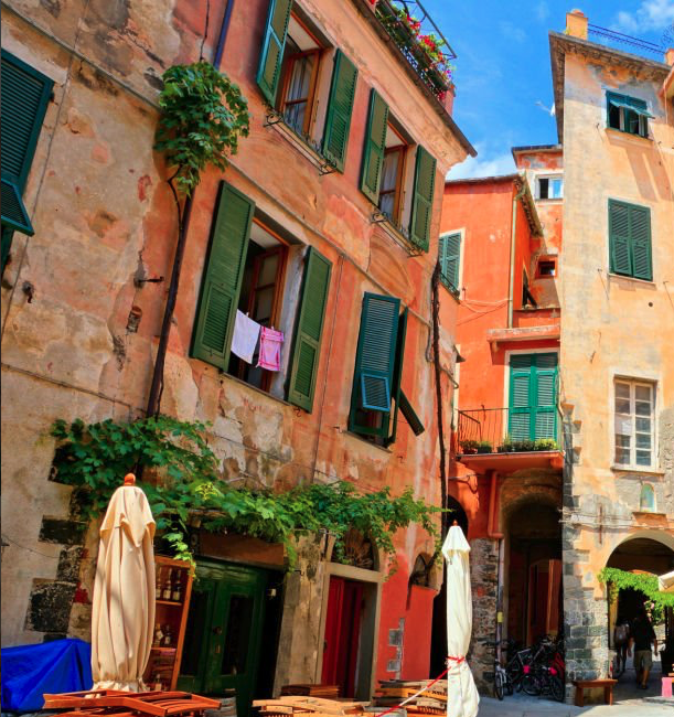 Monterosso al mare with colorful buildings with green shutters, laundry hanging outside the window, and outdoor seating with tables and umbrellas on a sunny day in an old European city street.