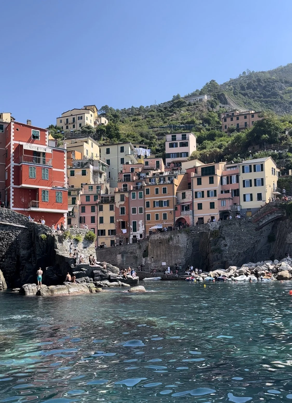 Riomaggiore with colorful buildings on a hillside next to turquoise water, with people swimming and relaxing on rocks and boats, under a clear blue sky.