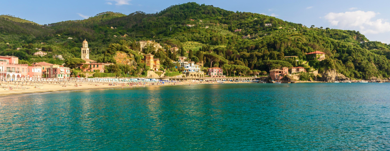 Levanto with colorful seaside town with pastel buildings on a beach, backed by green hills and mountains under a partly cloudy sky.