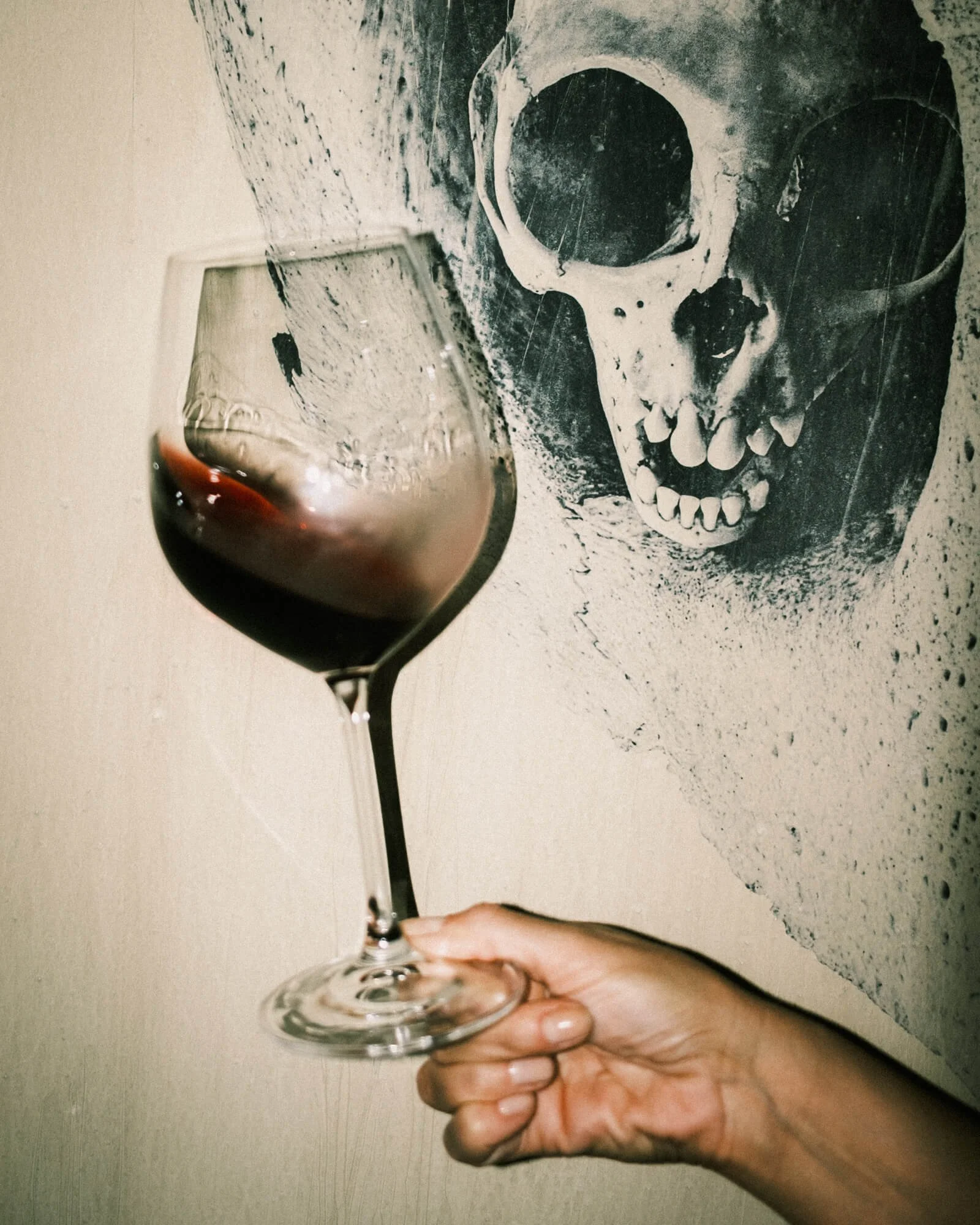 A hand holding a glass of red wine in front of a wall with a black and white skull painting.