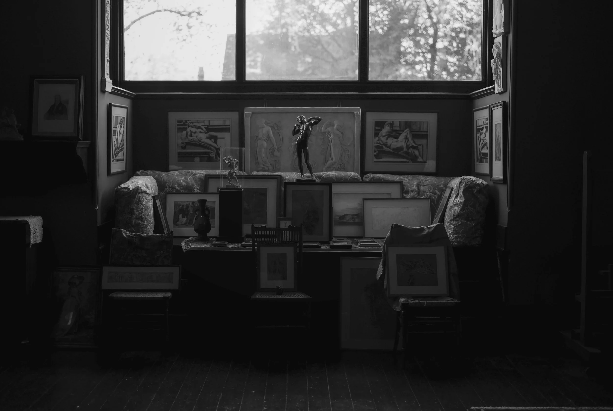 An art gallery with numerous framed paintings and sculptures arranged on a sofa and gallery table, illuminated by natural light coming from a large window.