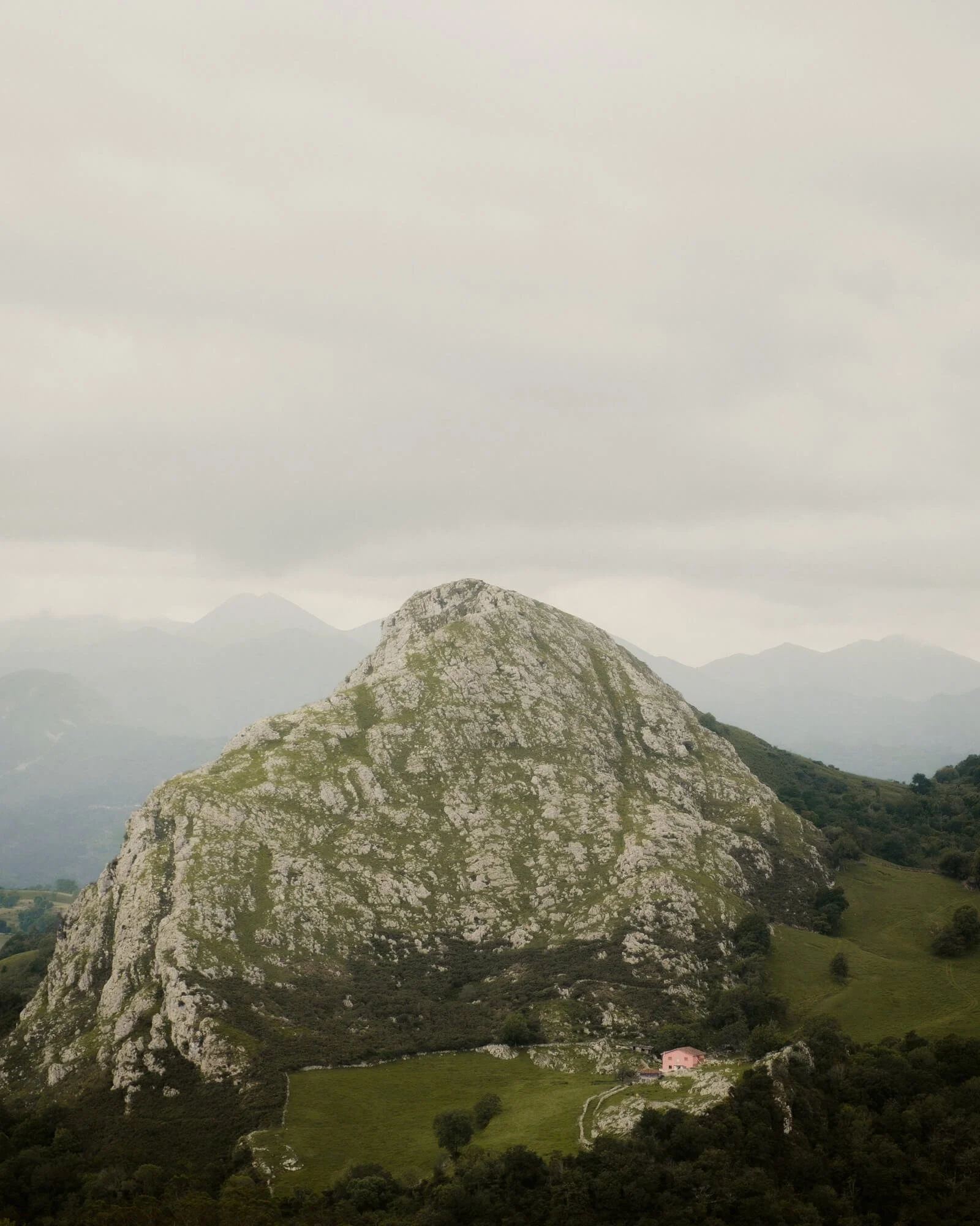 A large, rocky mountain with a steep, rounded top surrounded by green hills and trees, with a small pink house at the base. Overcast sky with gray clouds.