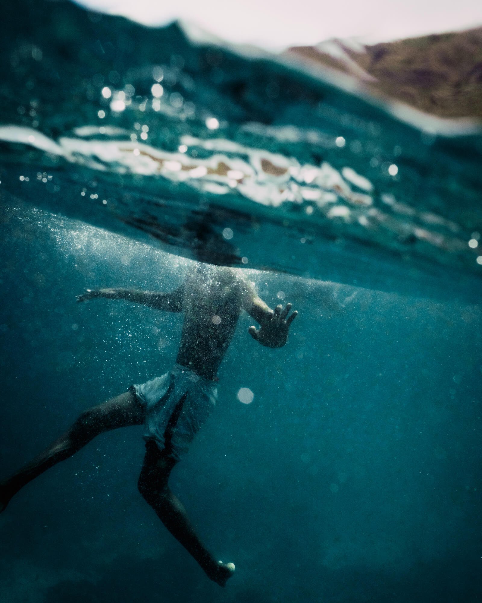 People swimming underwater in the ocean or a pool, with waves and mountains in the distant background.