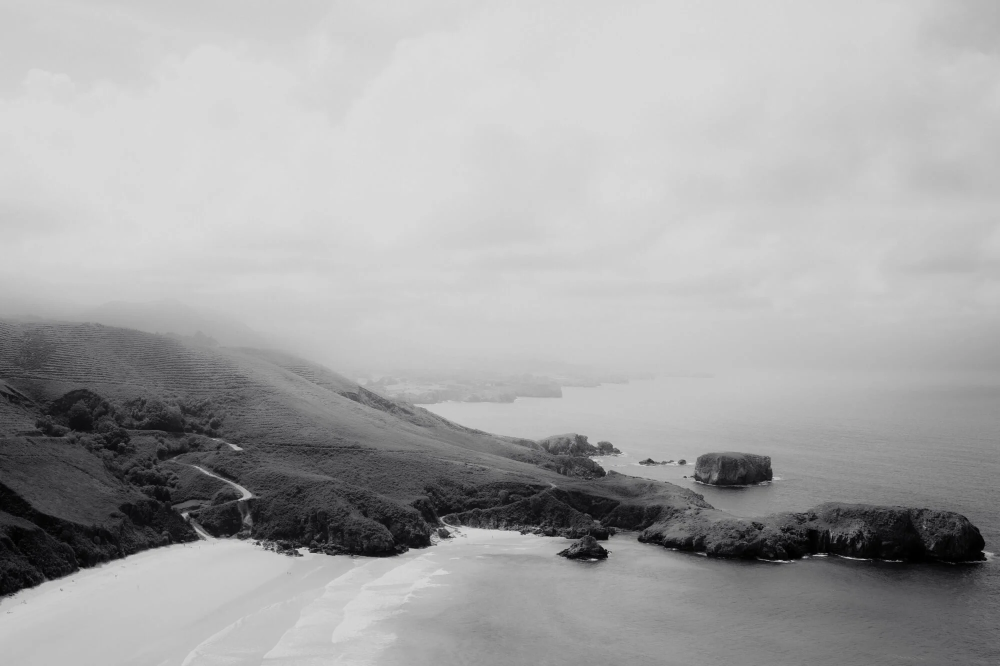 Black and white landscape of a coastal hillside with winding paths, cliffs, and the ocean in the background, under cloudy skies.