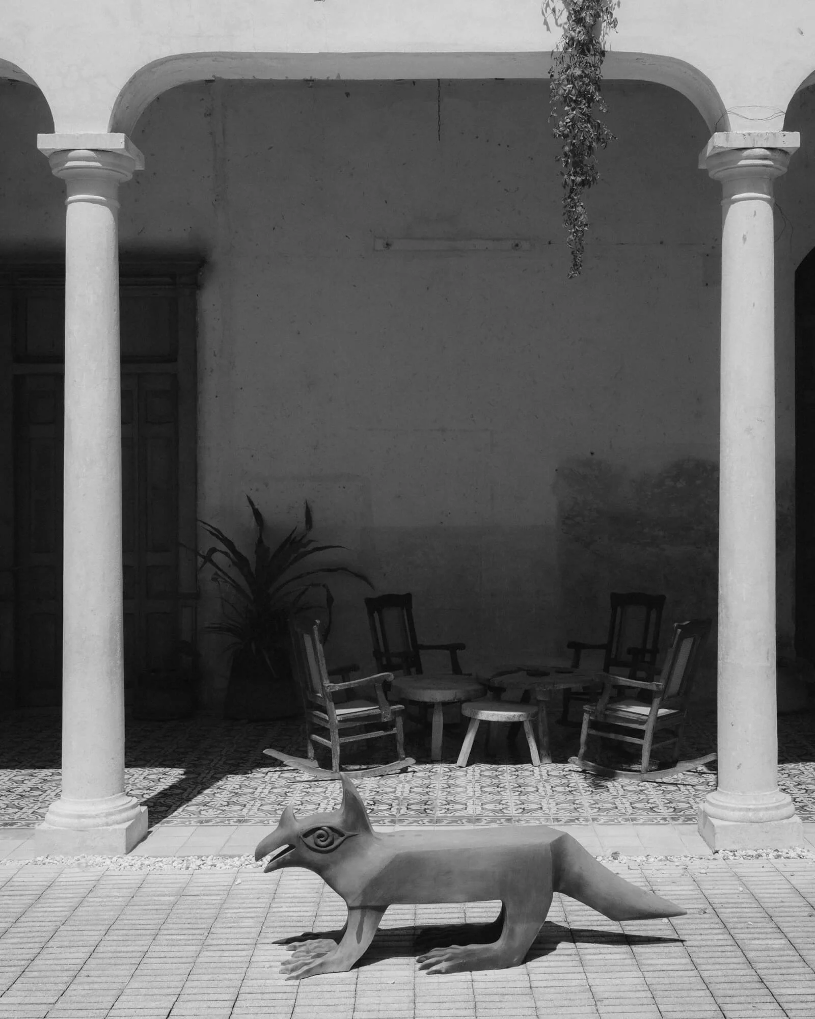 Black and white photo of a courtyard with a seating area, featuring a metal sculpture of a animal with a long neck and head, sitting on the tiled floor. Two columns support an archway, and there is a hanging plant and some chairs and a table in the background.