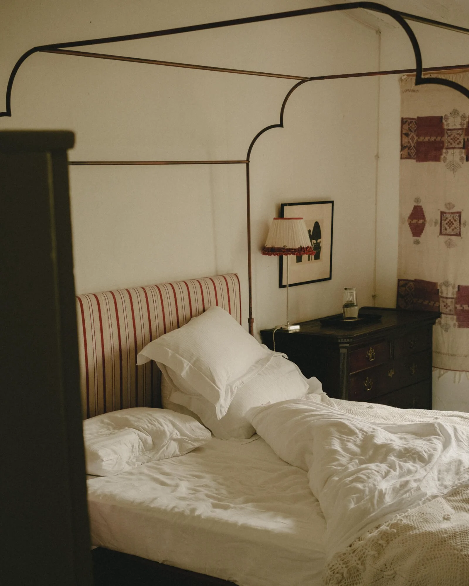 Unmade bed with white linens, striped and patterned headboard, bedside table with lamp, framed artwork, water glass, and tapestry on the wall.