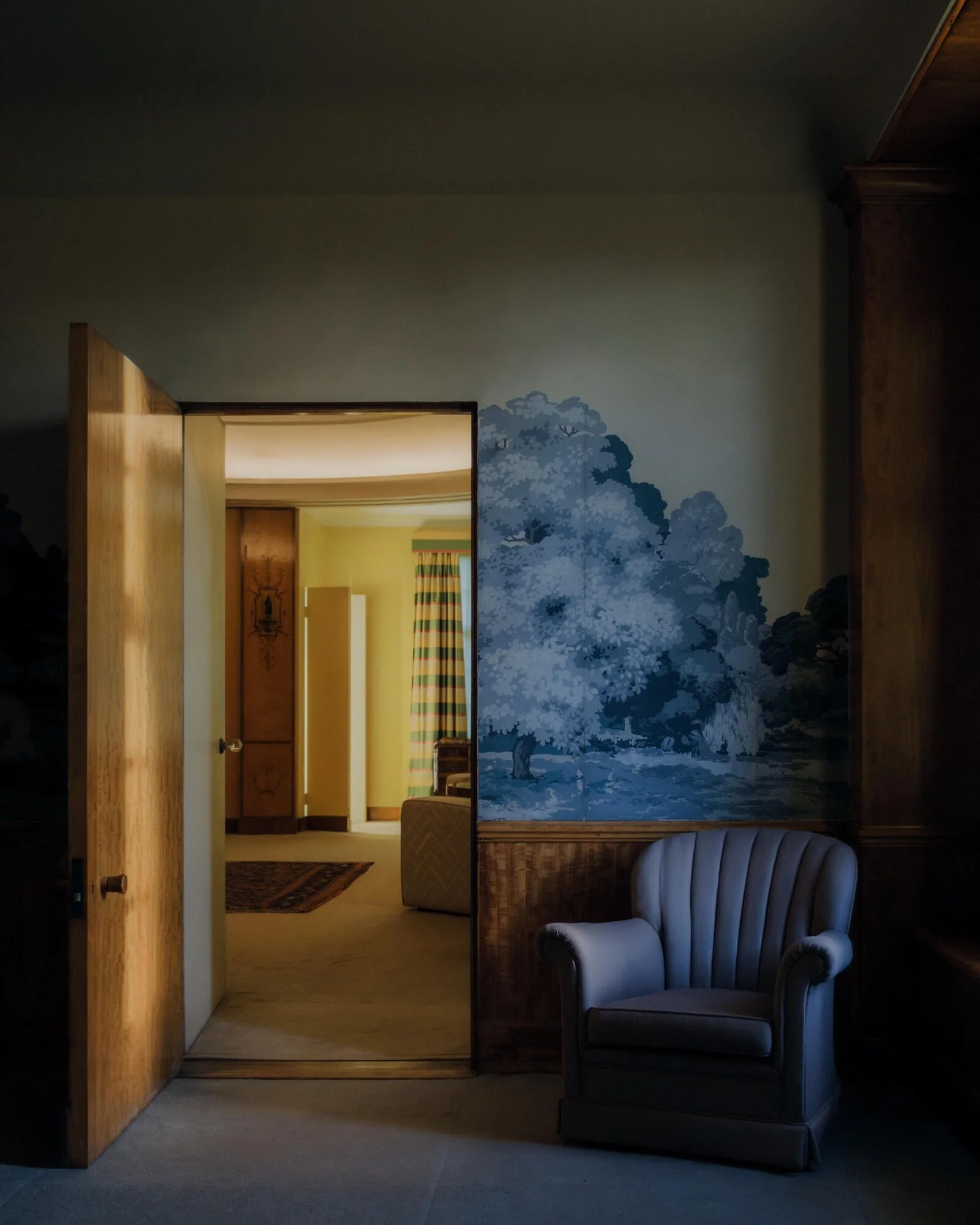 A room with a wall mural of a large tree and sailboats, a wooden door open to a hallway with a yellow wall and a painting, a beige armchair, and a striped window curtain.