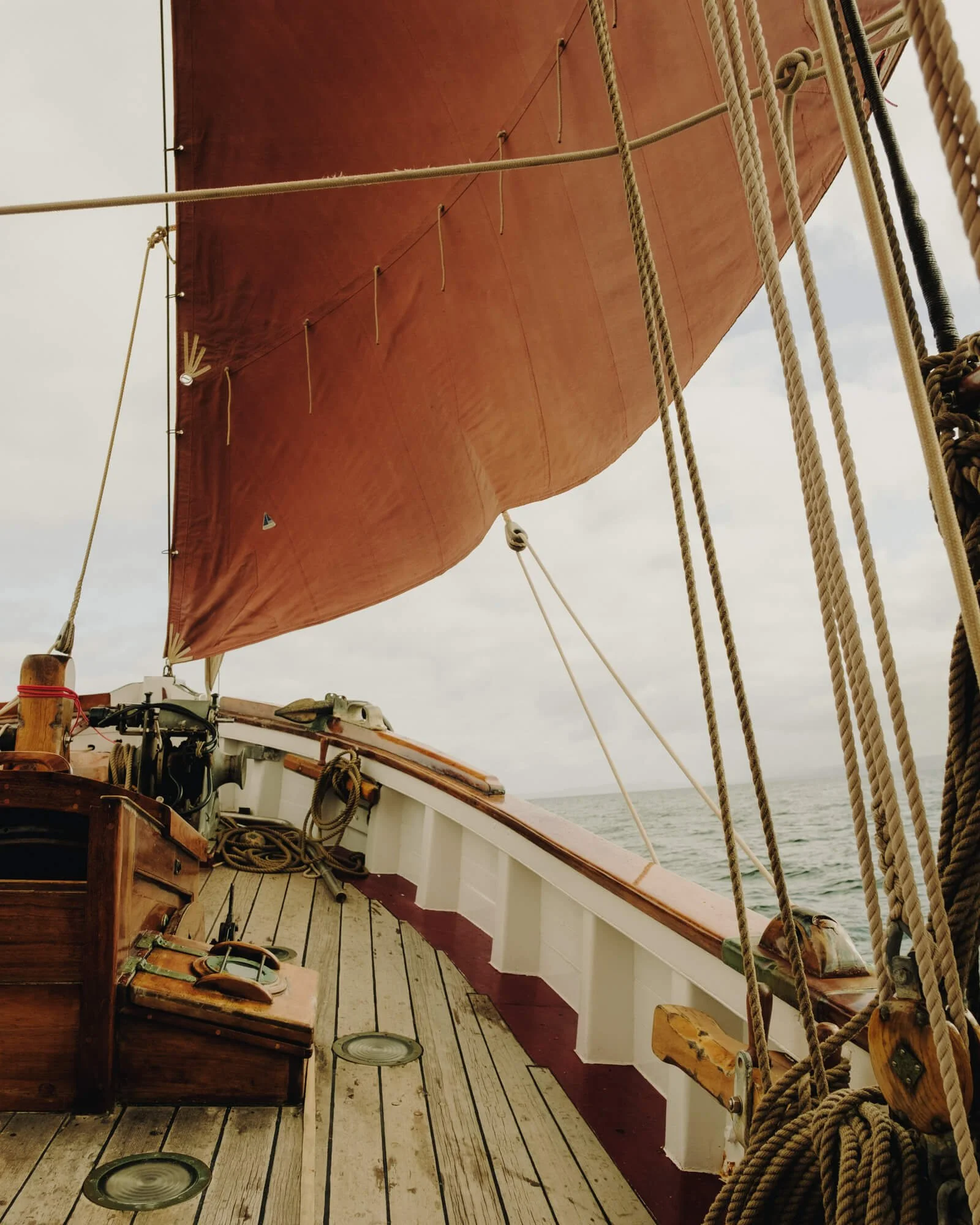 View of the deck of a wooden sailing ship with rigging and ropes, and a large, angled red sail against a cloudy sky and open water.