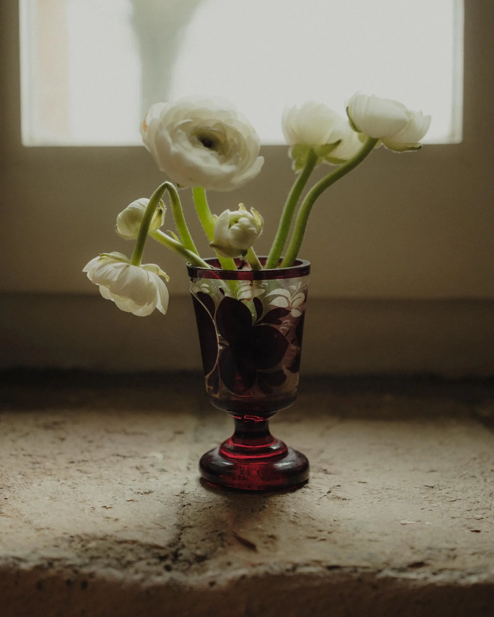 A bouquet of white flowers in a red and black decorative glass vase, placed on a light-colored textured surface, with a plain white background.