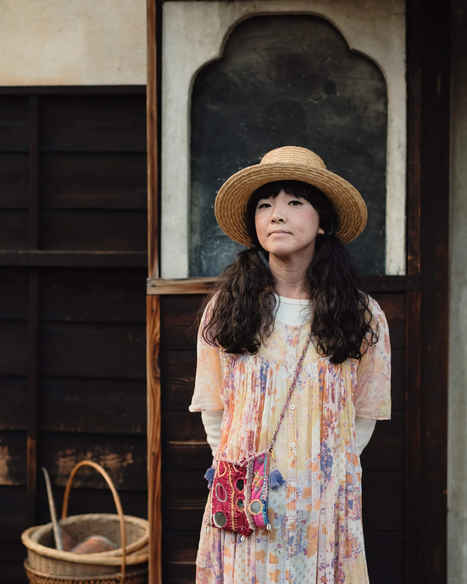 A woman with long dark hair wearing a wide-brimmed straw hat and a colorful, patterned dress standing in front of a dark wooden wall with a small window or opening behind her.