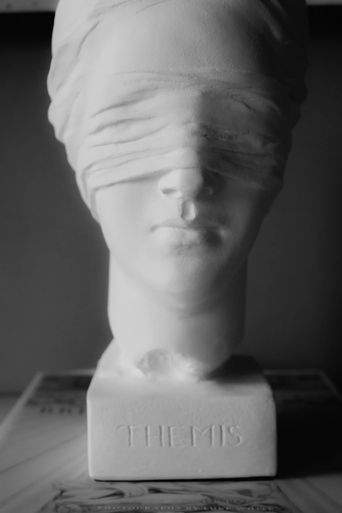 A white bust of a woman's face with closed eyes and lips, wrapped in flowing bands, on a rectangular base with the inscription 'TITUS' and partially visible 'TIMES'.