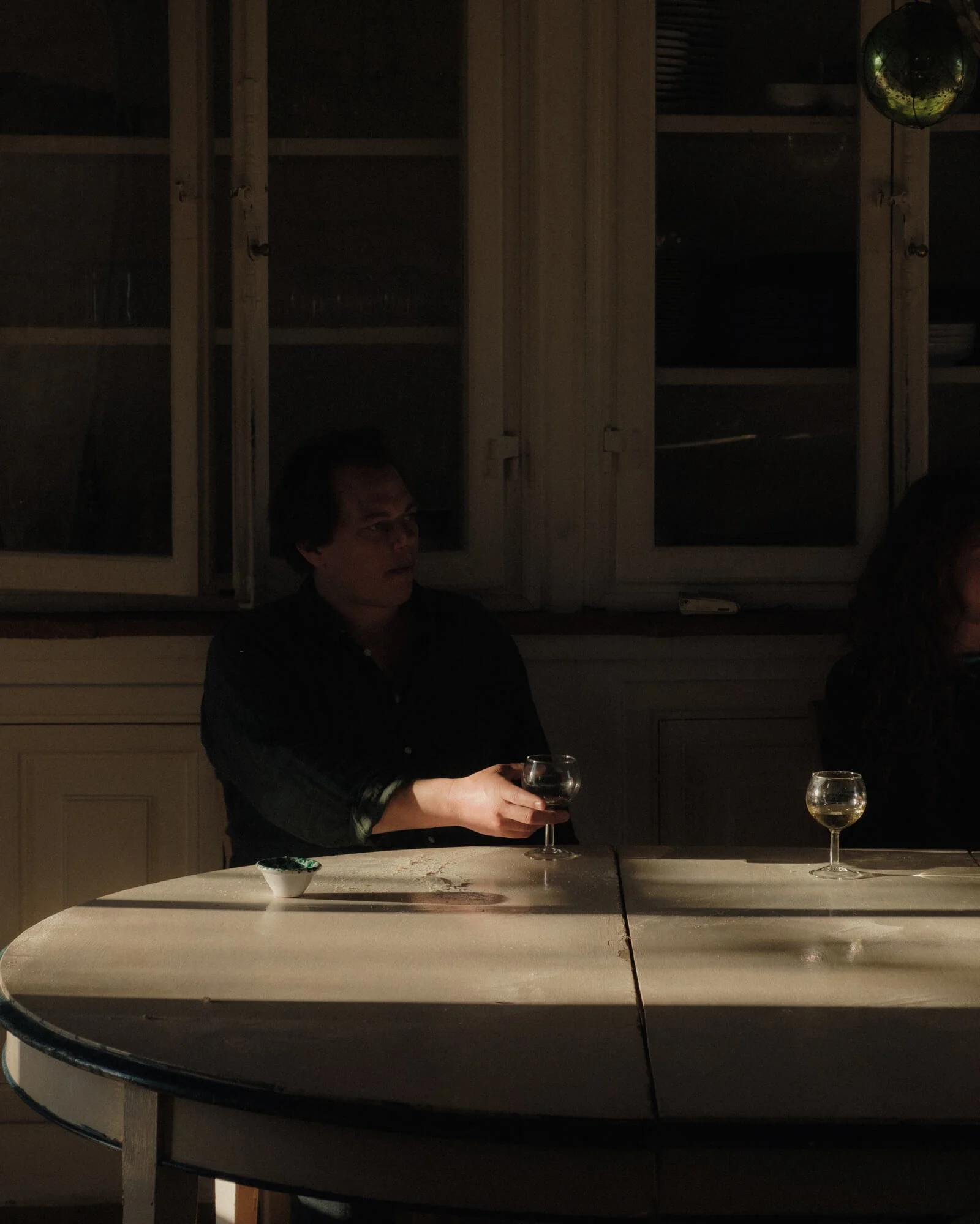 A person sitting at a dining table holding a glass of wine in a dimly lit room with wooden cabinets and another person partially visible to the right.