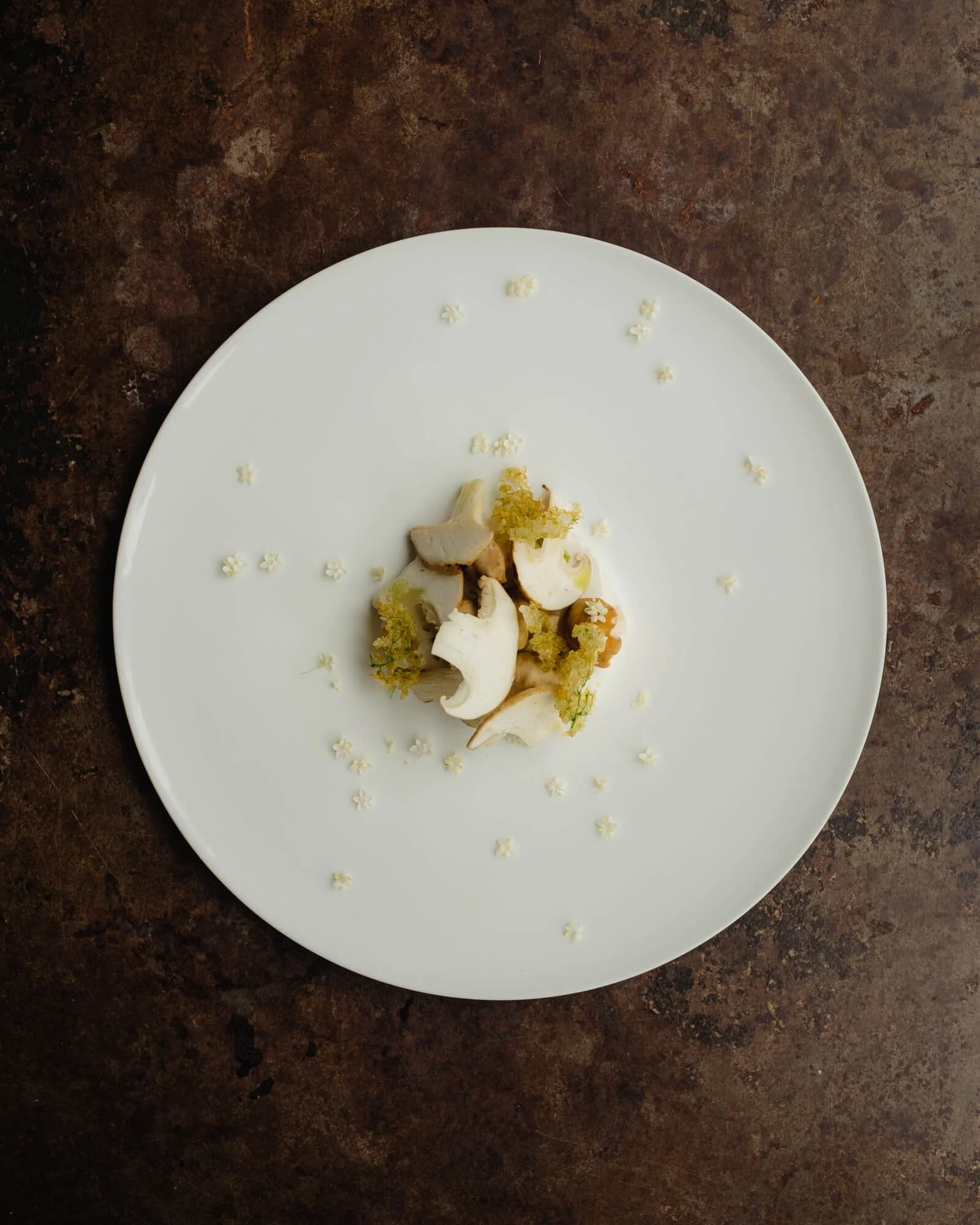 A white plate with small decorative flowers around the edge, holding a small pile of peeled and sliced vegetables, on a dark brown textured surface.