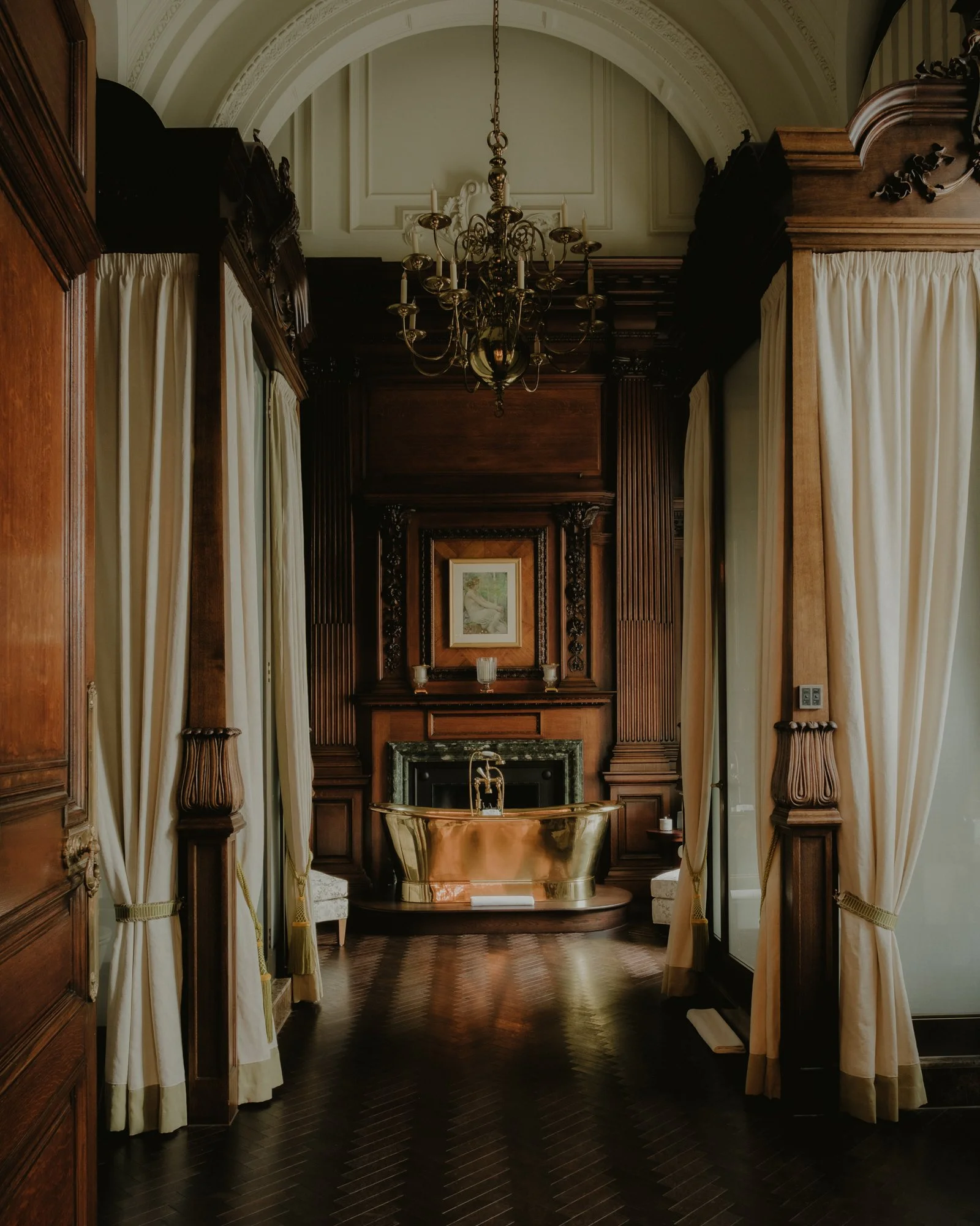 Elegant bathroom with dark wood paneling, a large gold bathtub, and cream-colored curtains framing the space. A chandelier hangs from the arched ceiling, and a framed painting is visible on the wall above the bathtub.