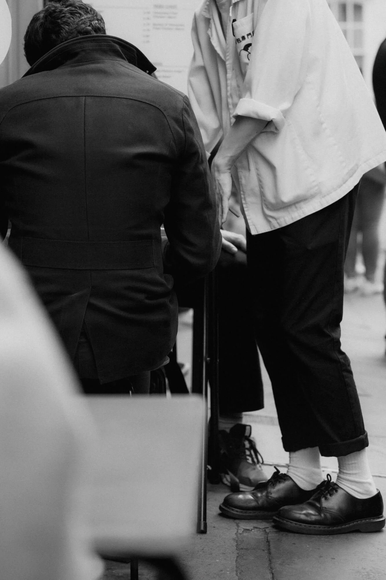 Black and white photo of two people standing at a counter, one person seated and the other leaning over, engaging in a transaction or conversation. The person standing is wearing a light-colored jacket, dark pants, socks, and shoes, while the seated person wears a darker jacket. The scene appears to be in an indoor setting, possibly a store or office.