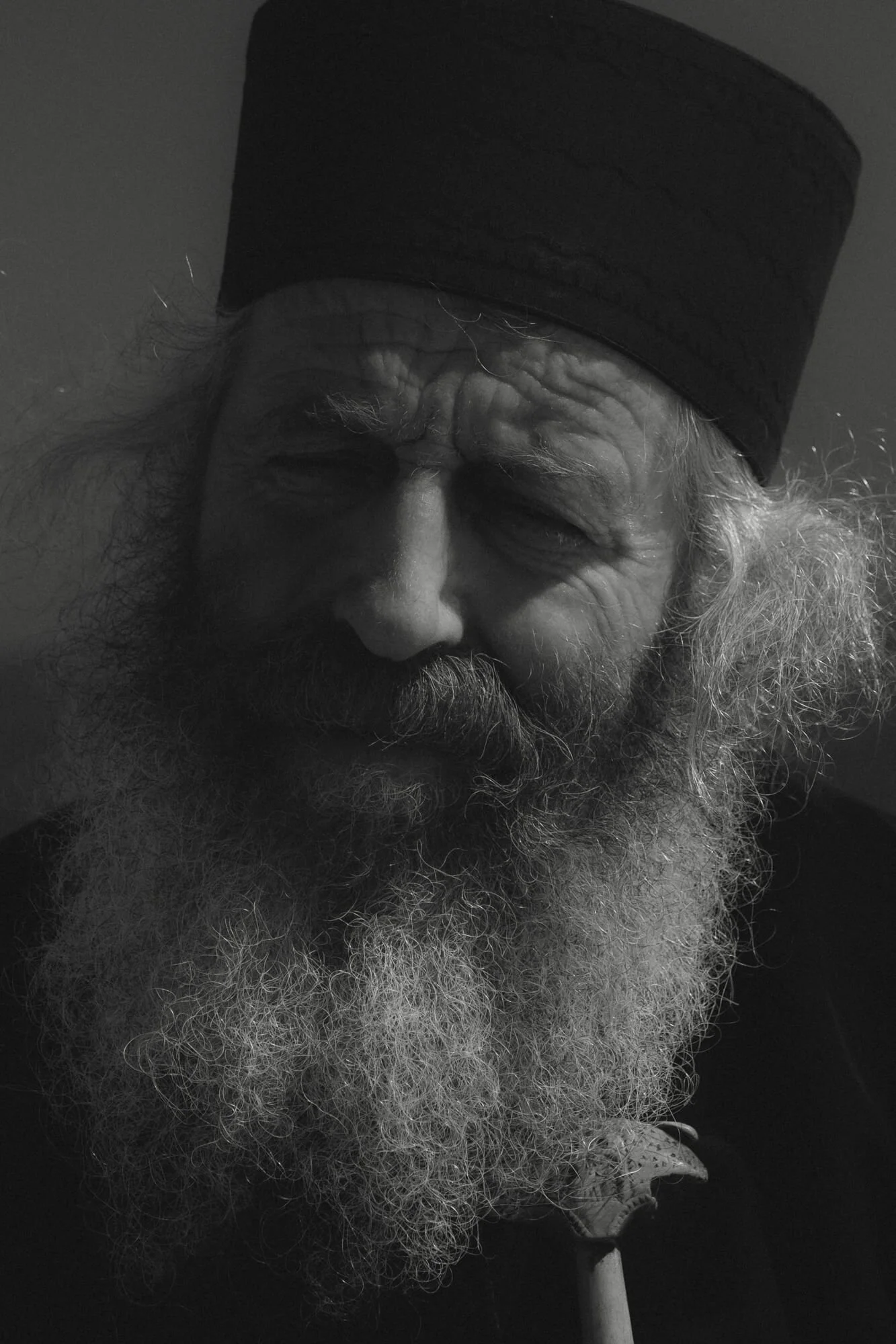 Black and white close-up photo of an elderly man with a long white beard, curly hair, wearing a black beanie, with a somber expression.