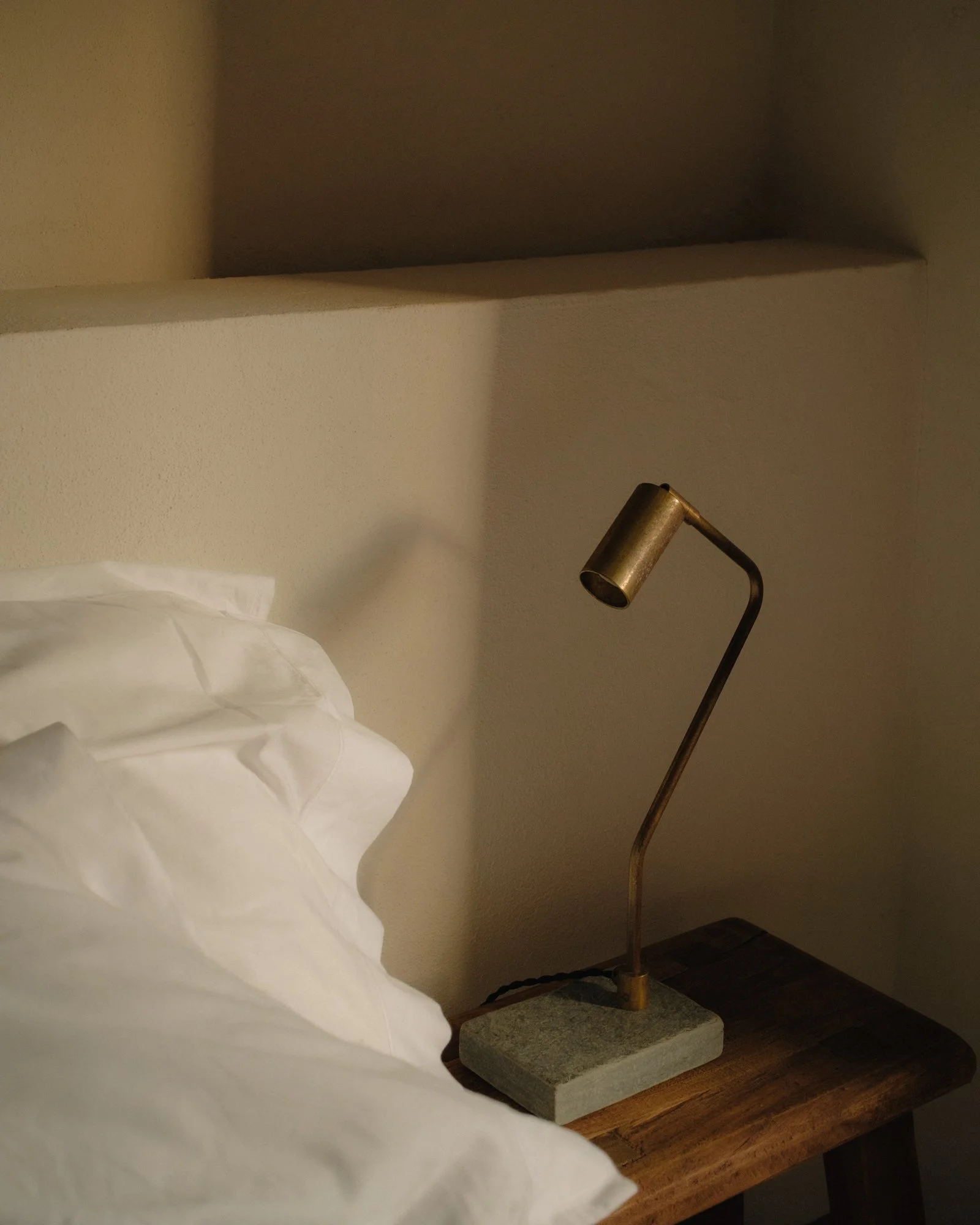 A brass-colored reading lamp on a wooden bedside table and a white pillow on a bed with beige walls.