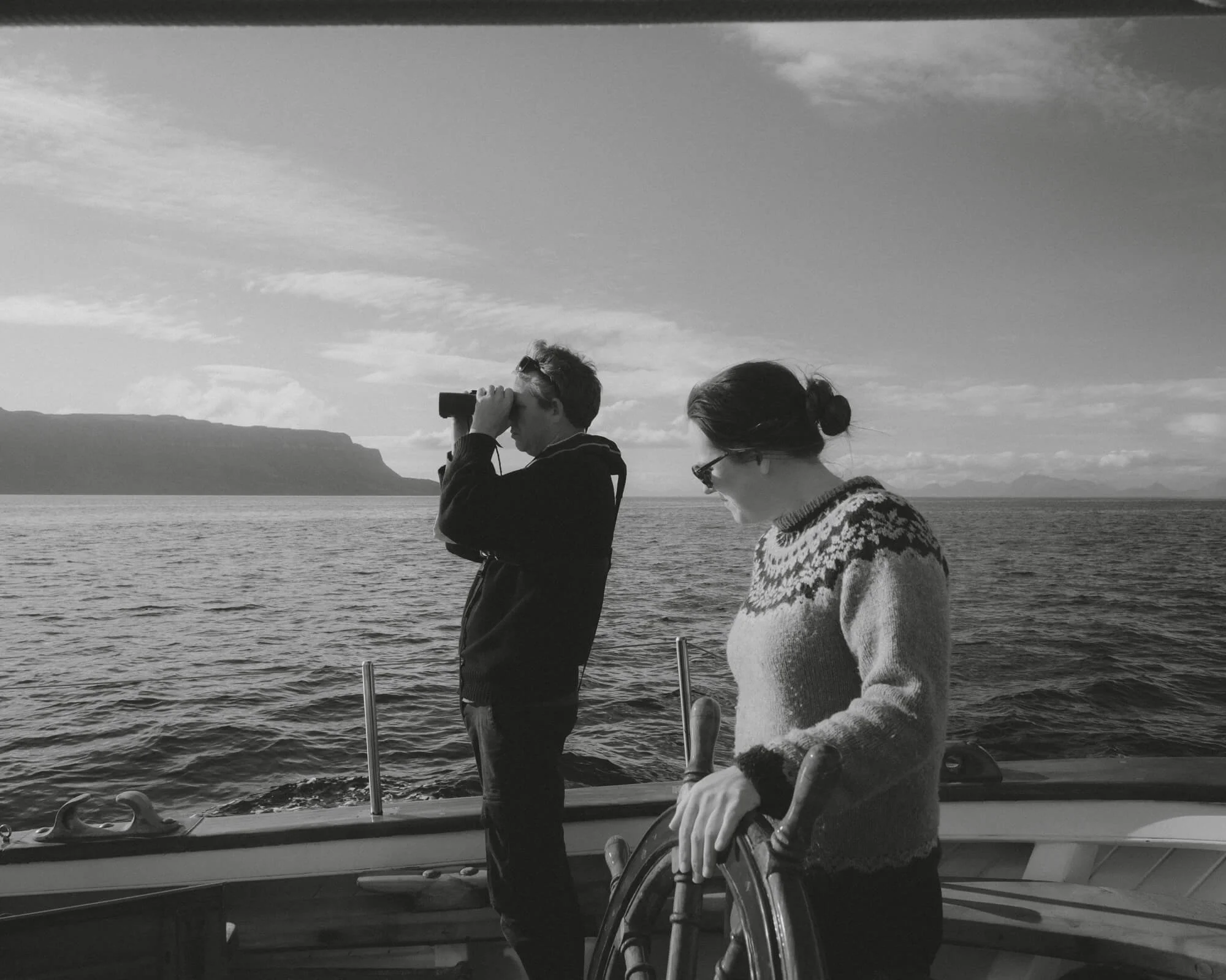Two people on a boat, one using binoculars and the other in a wheelchair, with water and a mountain in the background.