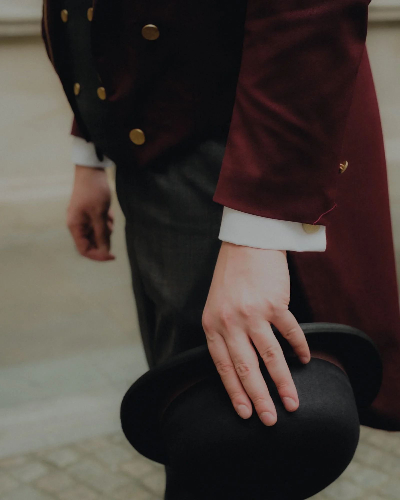 Close-up of a person's hand resting on their lap, wearing dark pants, a white shirt, and a maroon blazer with gold buttons.