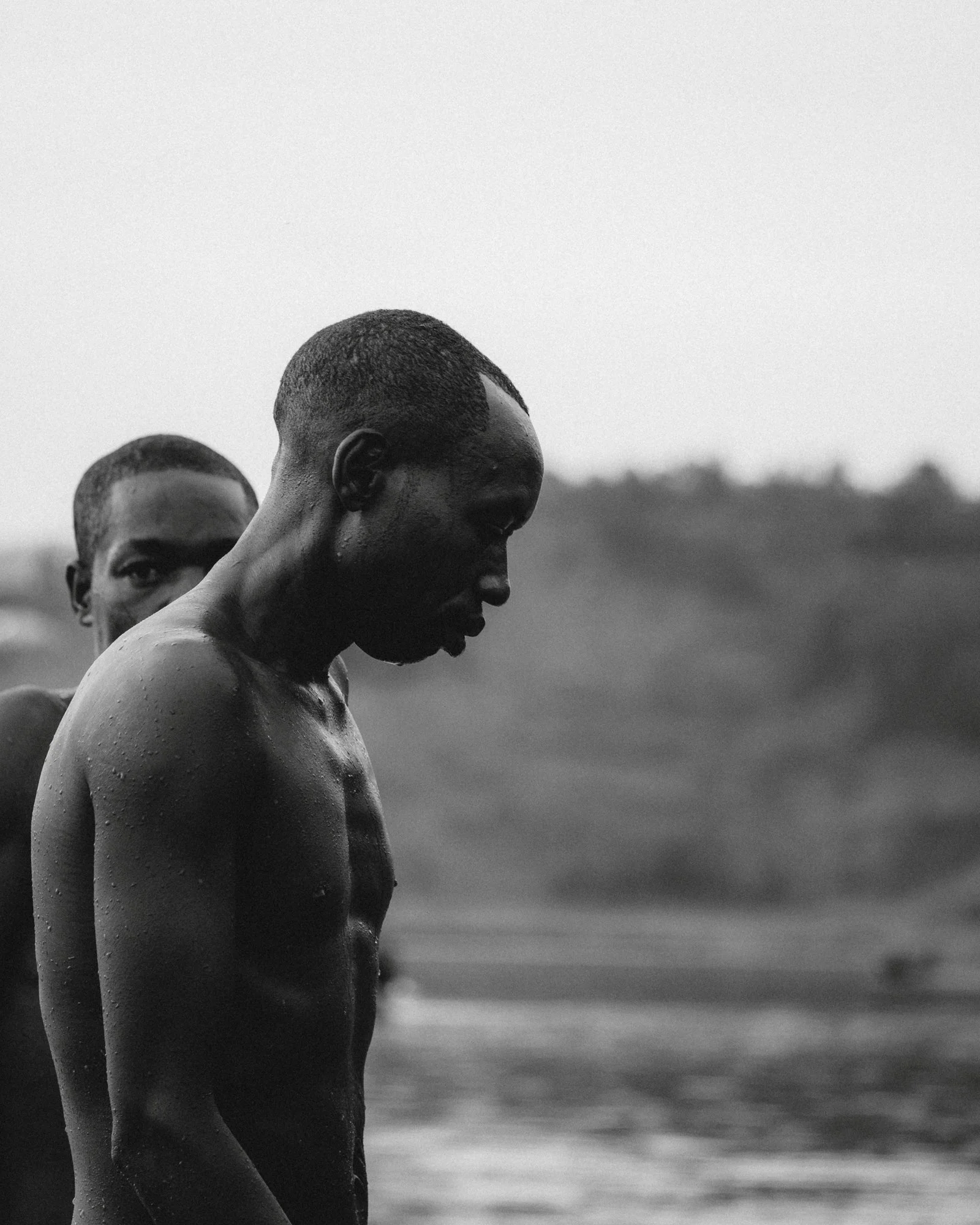 Close-up of two dark-skinned, shirtless young men with short, wet hair, standing outdoors in profile, with a blurred natural landscape background.
