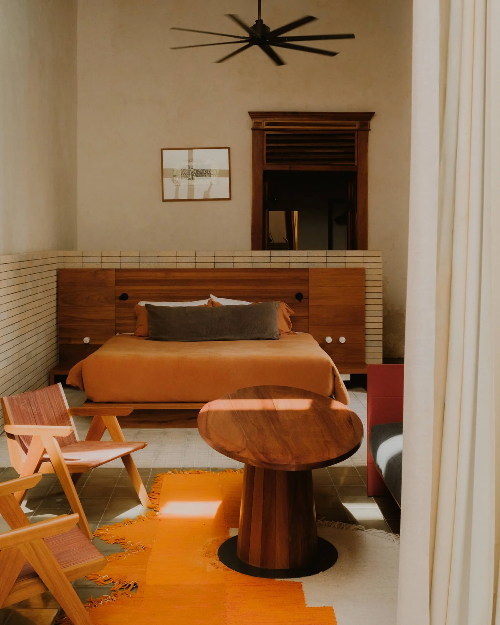 A cozy bedroom with a wooden bed, orange duvet, and gray pillow, illuminated by sunlight, with modern furniture and decor, including a wooden table, chairs, a round mirror, and framed artwork on the wall.