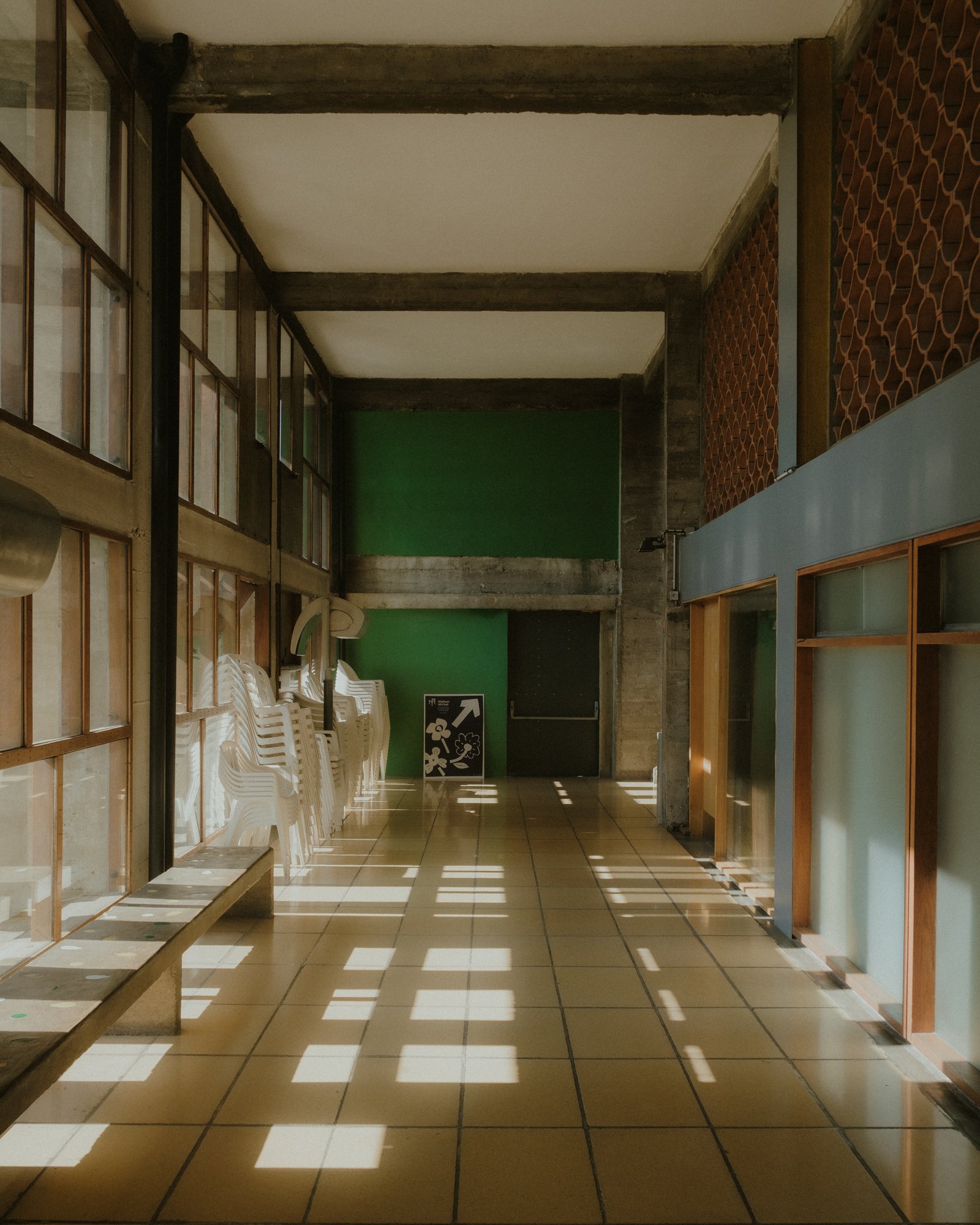Empty hallway with white chairs stacked on the left, green wall in the distance, and sunlight casting square-shaped shadows on the tiled floor.