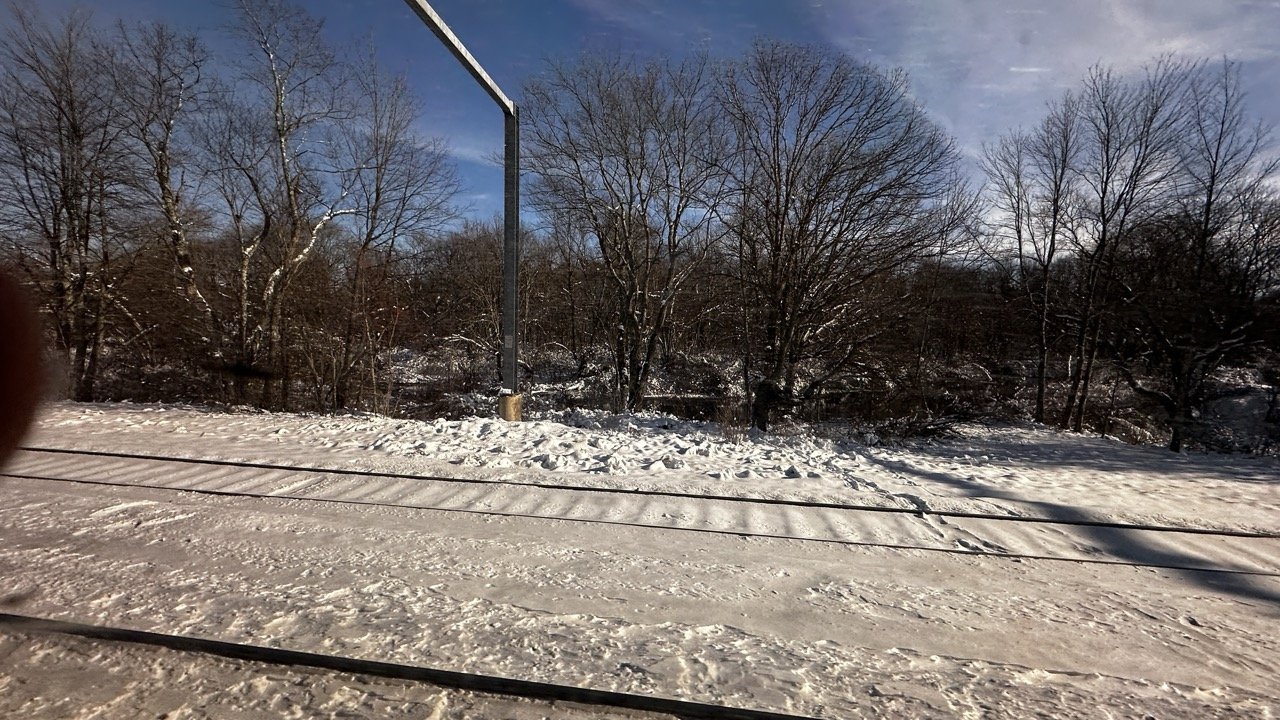 Snow-covered train tracks and a winter landscape with bare trees, viewed from an Amtrak window.