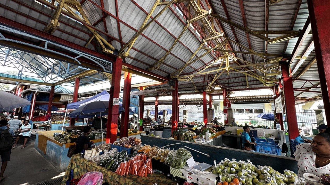 A bustling market scene in Victoria, Seychelles, with stalls selling fresh produce, vegetables, and fruit under a traditional tin roof.